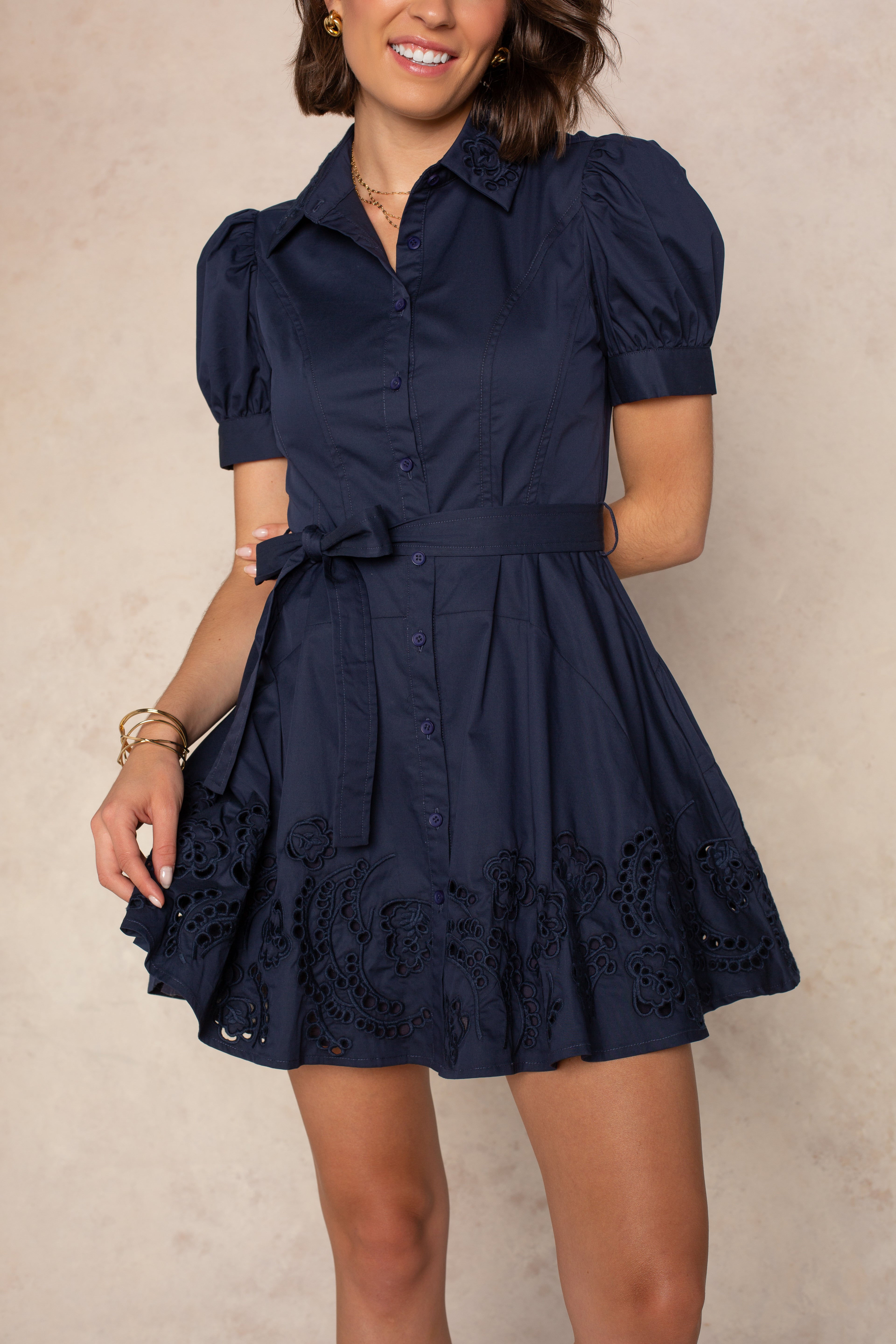 A woman wears the Georgie Madison Short Sleeve Mini Shirtdress in NAVY, featuring puff sleeves, a belted waist, and an eyelet embroidery hem. She accessorizes with bracelets and stands against a light, neutral background.