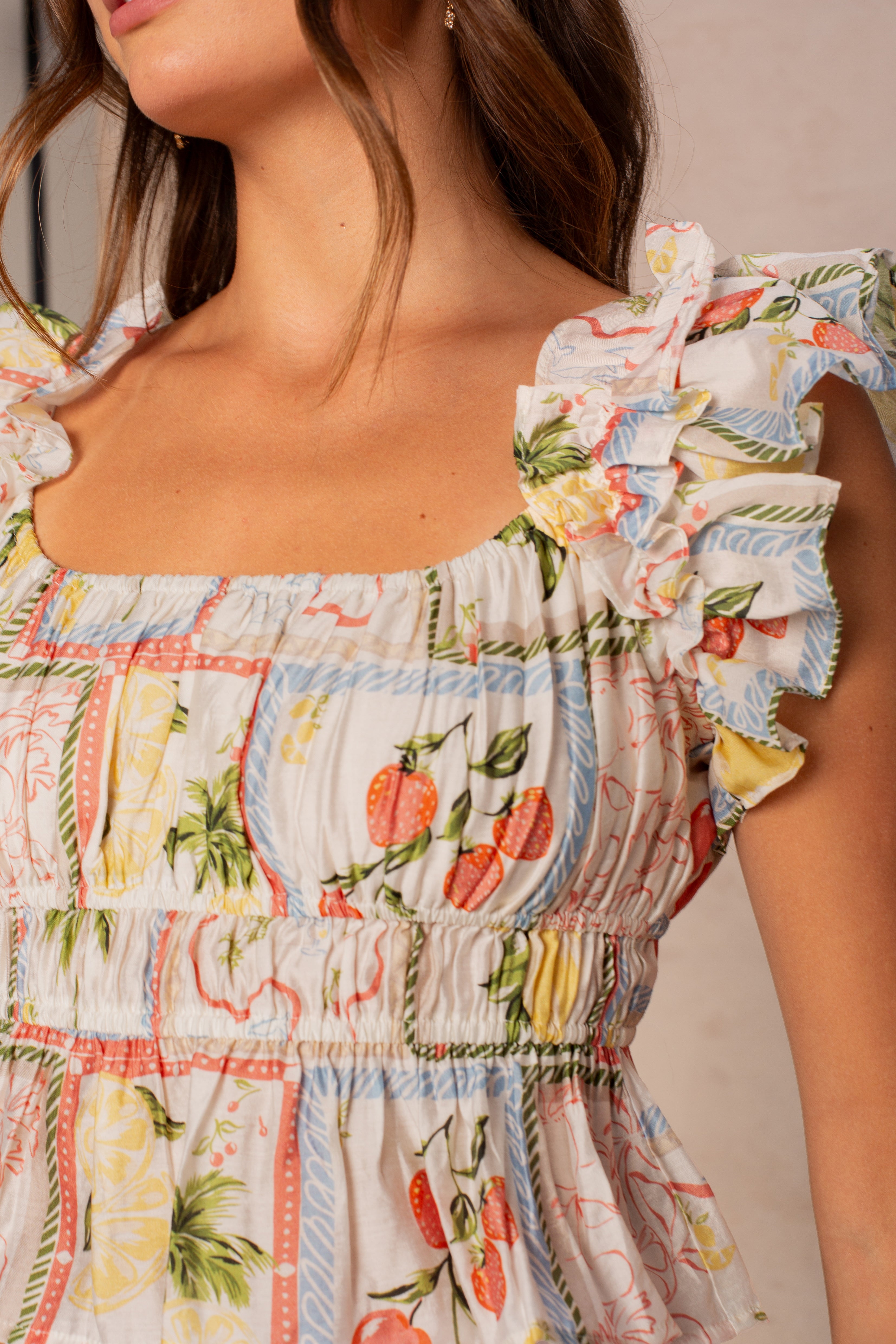 A woman wears the Georgie Avery Top In Multi, a ruffled, sleeveless top with a colorful fruit and floral pattern. The smocked top is adorned with strawberries, lemons, and leaves. Only her torso and part of her face are visible.