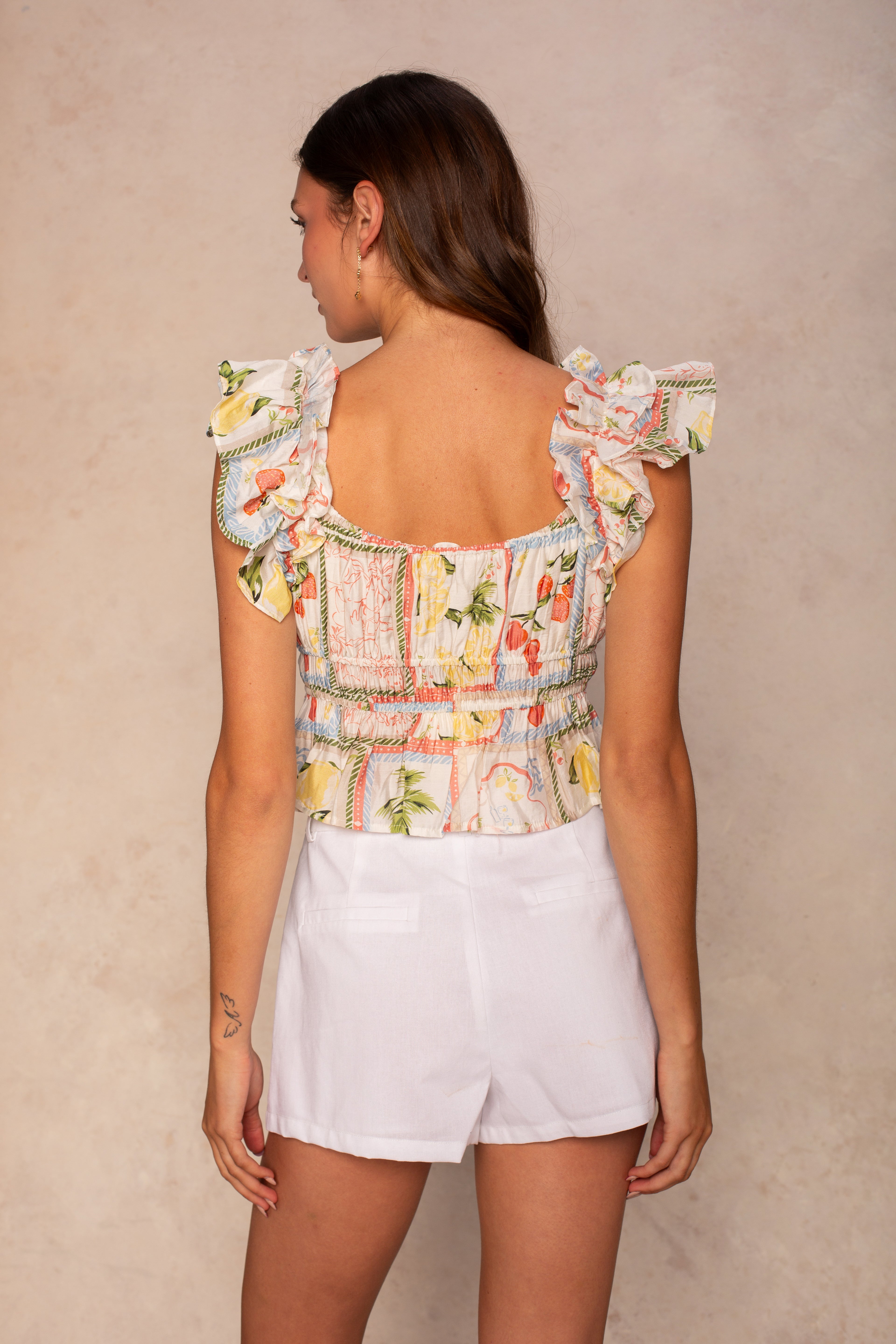 A woman with long brown hair stands with her back to the camera, wearing the Georgie Avery Top—a ruffled, tropical print sleeveless peplum hem top—paired with white shorts against a beige background.
