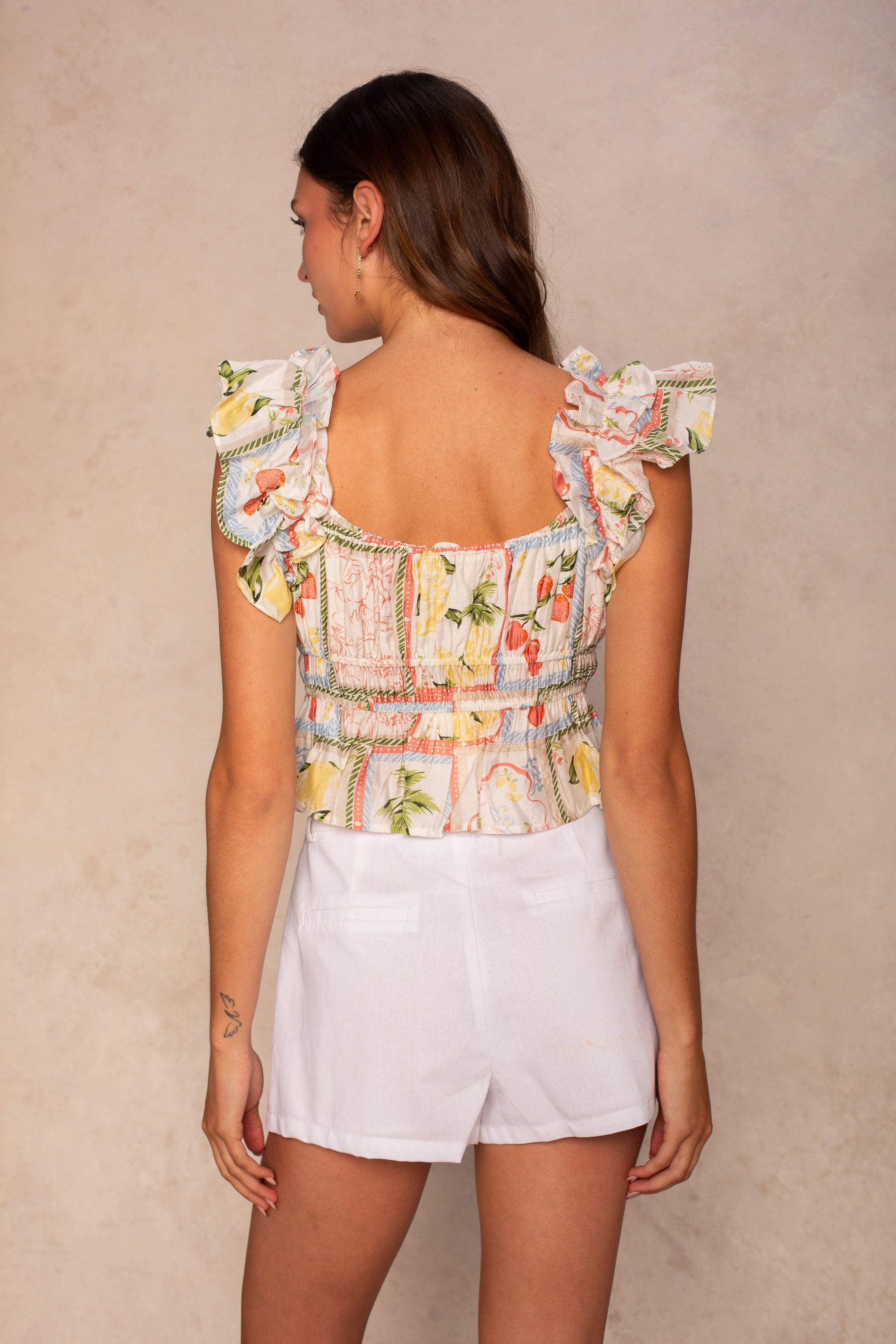 A woman stands with her back to the camera, wearing the Georgie Avery Top in Multi—a frilled, smocked top featuring a floral-patterned peplum hem—paired with white shorts against a beige background.
