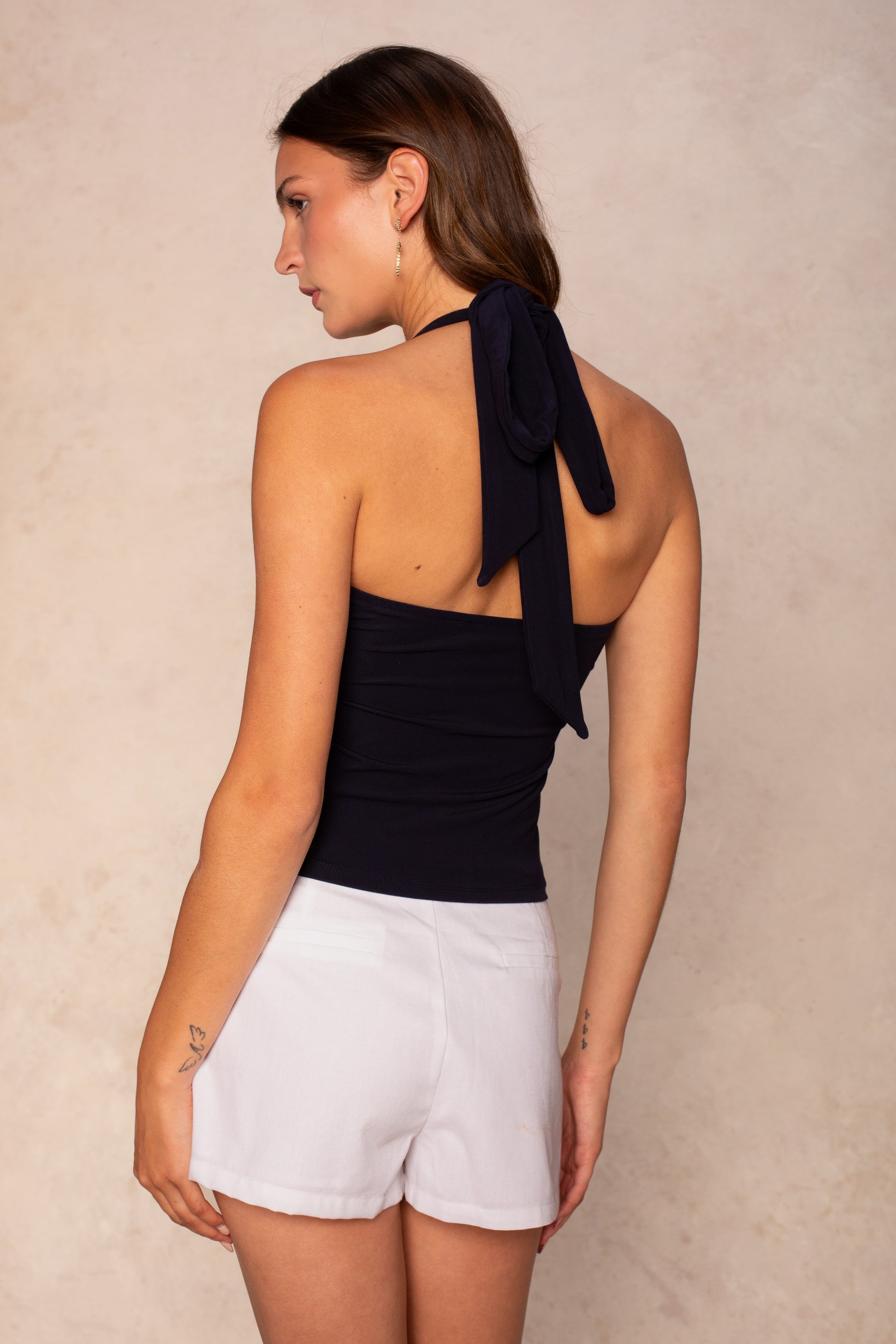 A woman with light skin and long brown hair wears the Georgie Julie Halter Tie Knit Top in navy, paired with white shorts and small hoop earrings, standing against a beige background.