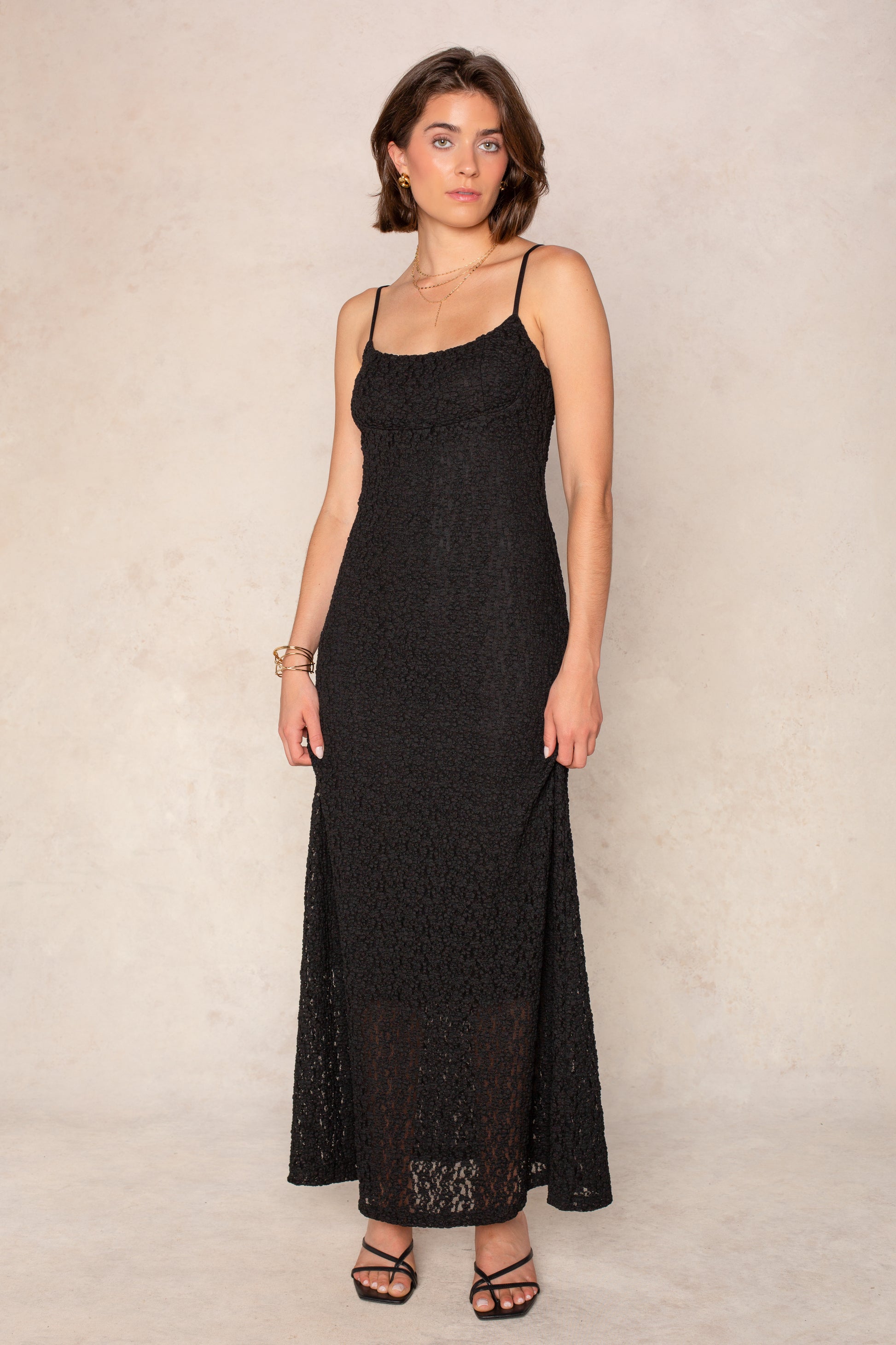 A woman in the Isla Reece Maxi Dress in BLACK, a sleeveless lace evening gown with thin straps, stands against a light textured background. She pairs it with black sandals, gold jewelry, and sports short brown hair for a timeless look.