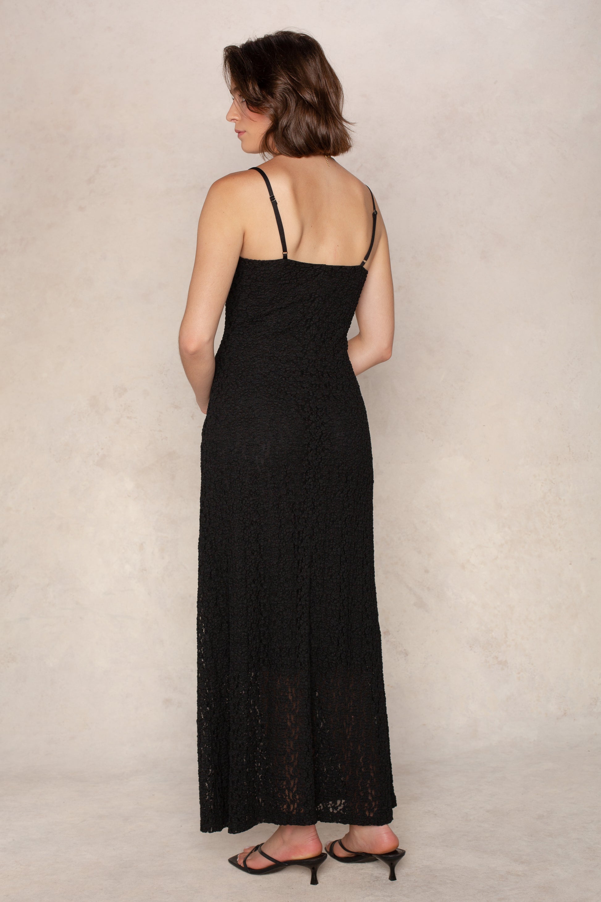A woman with short brown hair faces away from the camera, wearing the Isla Reece Maxi Dress—sleeveless, black, with thin straps—paired with black heeled sandals against a light textured backdrop.