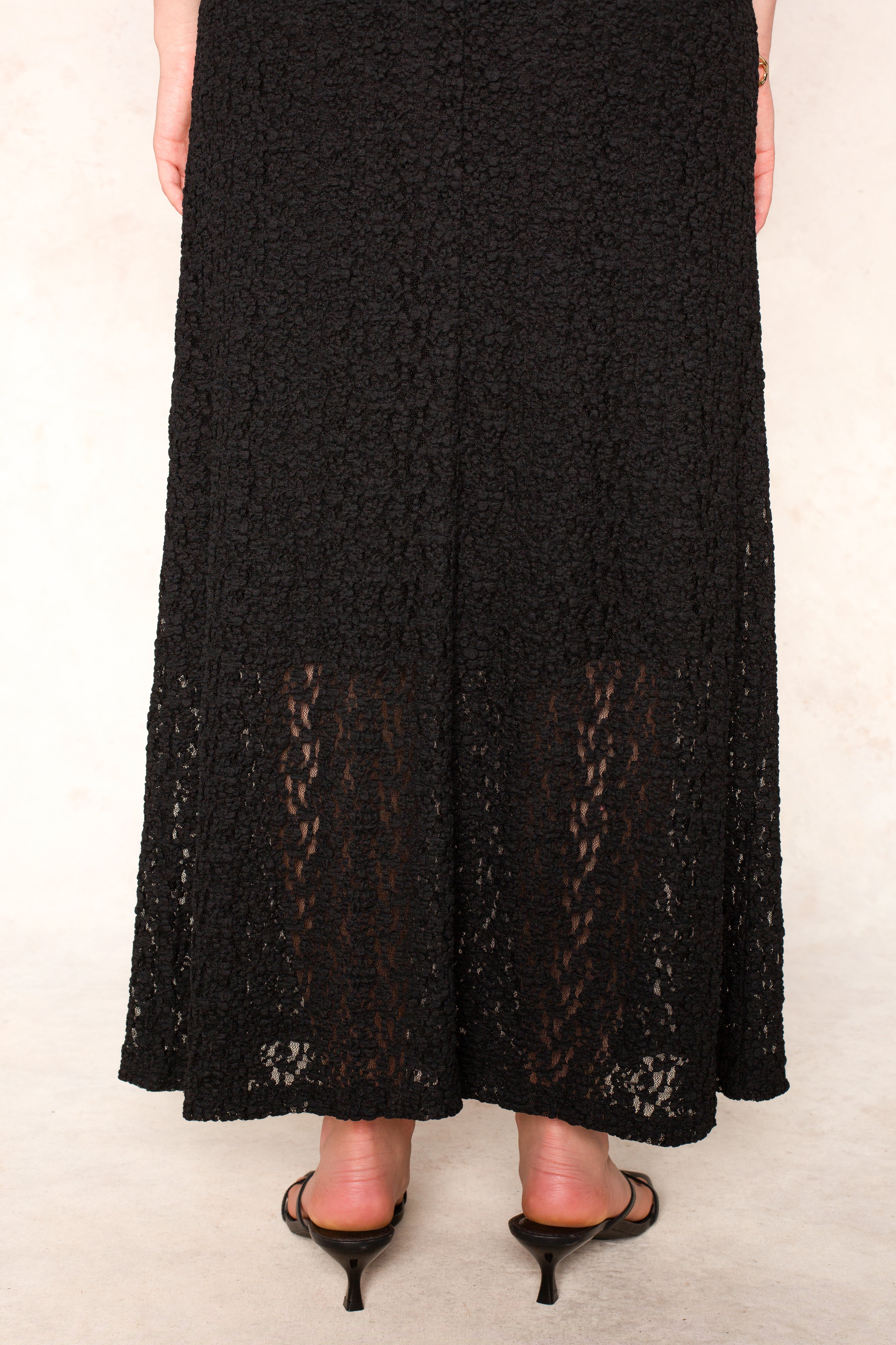 A person wears the Isla Reece Maxi Dress, a long textured black lace dress with a semi-sheer hem section, paired with black open-toe heels on a light floor.