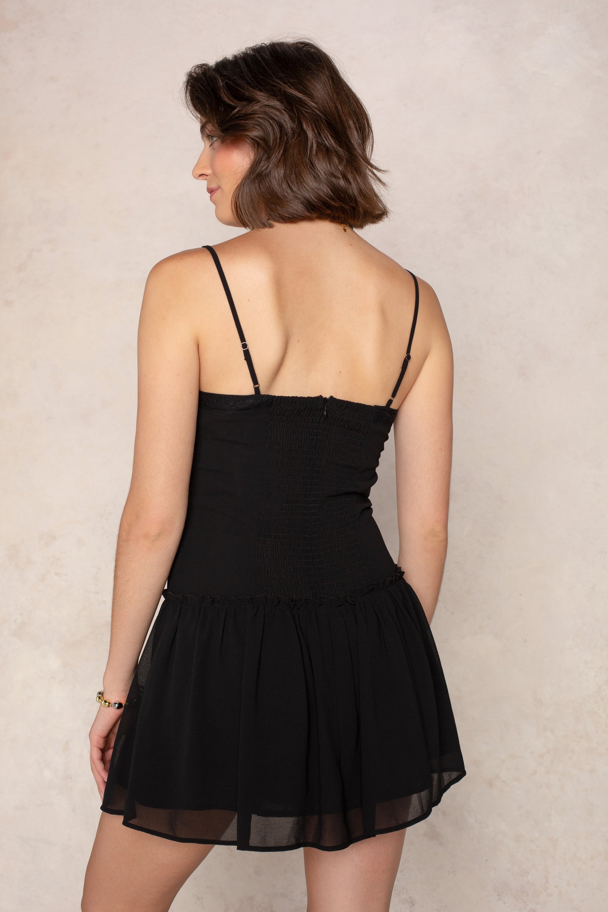 A woman with short brown hair stands with her back to the camera against a light, textured background, wearing the Isla Lottie Mini Dress—a sleeveless black dress featuring thin straps.