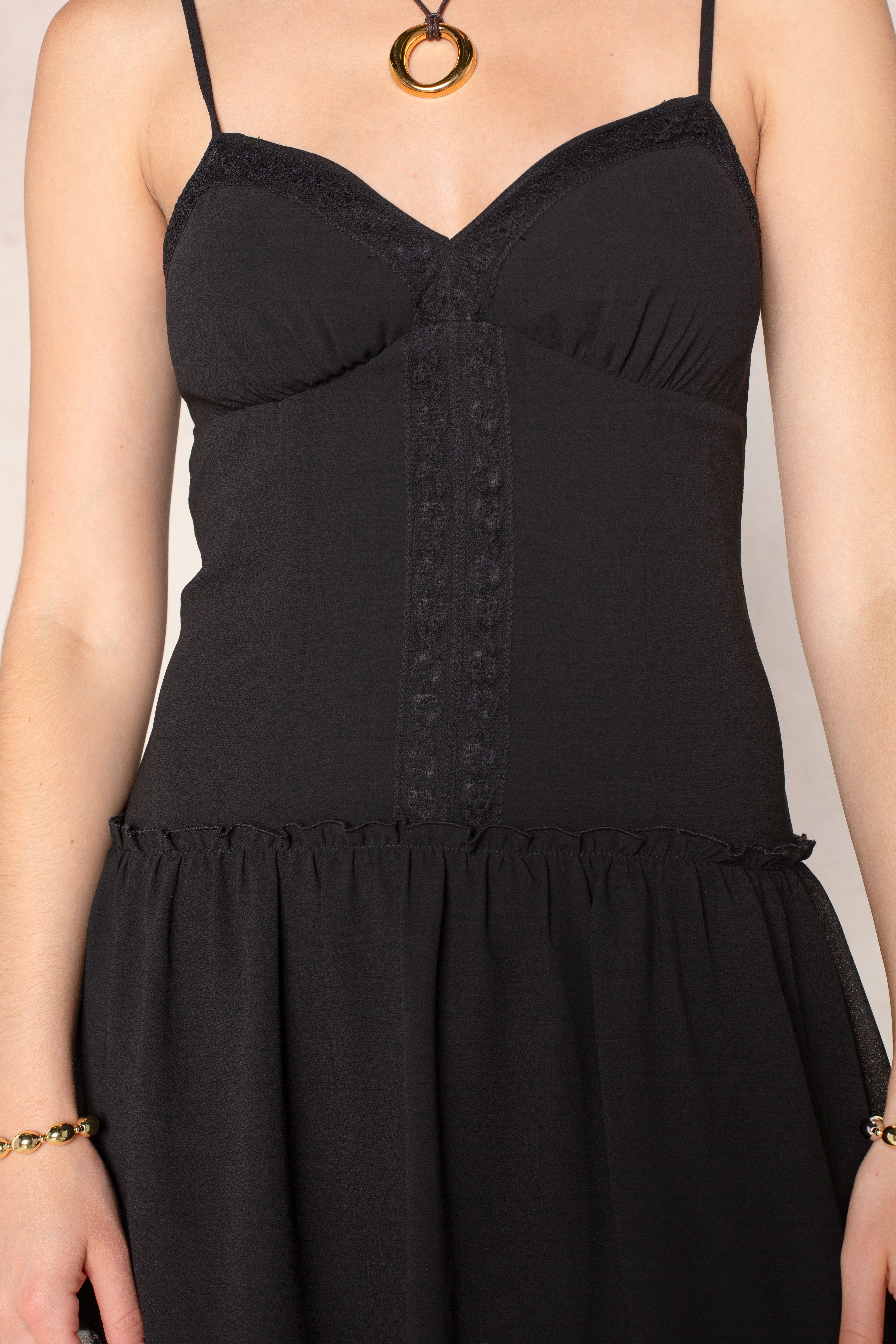 A person wearing the Isla Lottie Mini Dress, a black mini dress with spaghetti straps and delicate lace trim details, paired with a gold necklace featuring a round pendant. Only the upper body is visible.