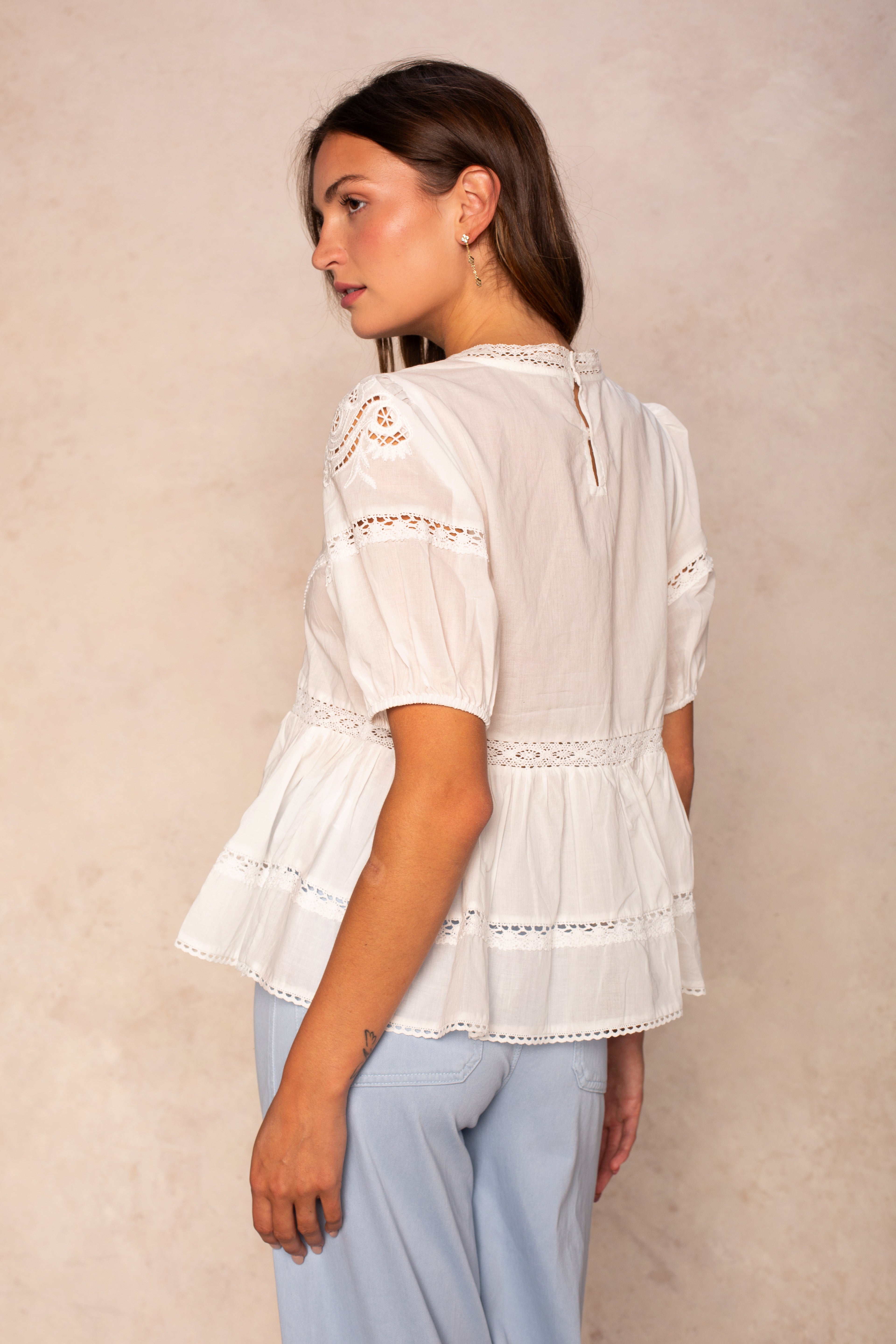 A woman stands sideways in the Georgie Rachel Eyelet Short Sleeve Blouse and light blue pants, posed against a beige textured background.