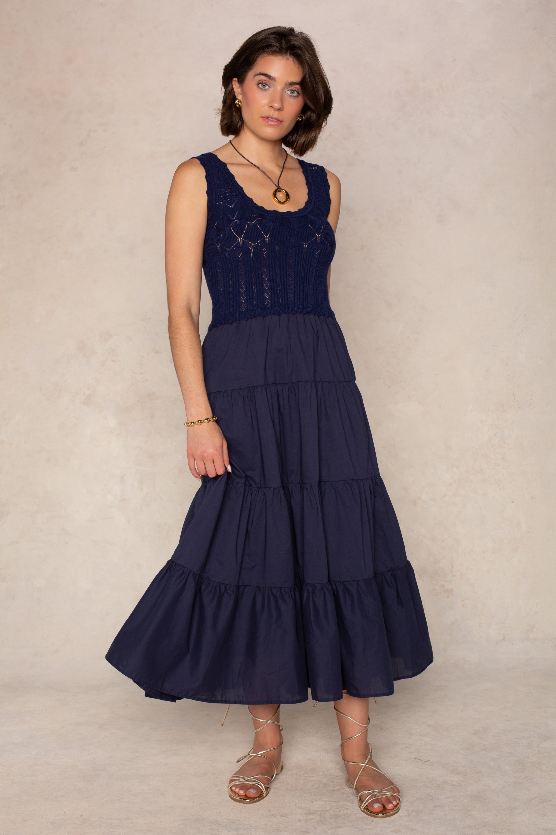 A woman stands against a neutral background wearing the Aria Midi Dress by Georgie in navy, featuring a fitted crochet bodice and tiered skirt, styled with gold jewelry and strappy sandals.