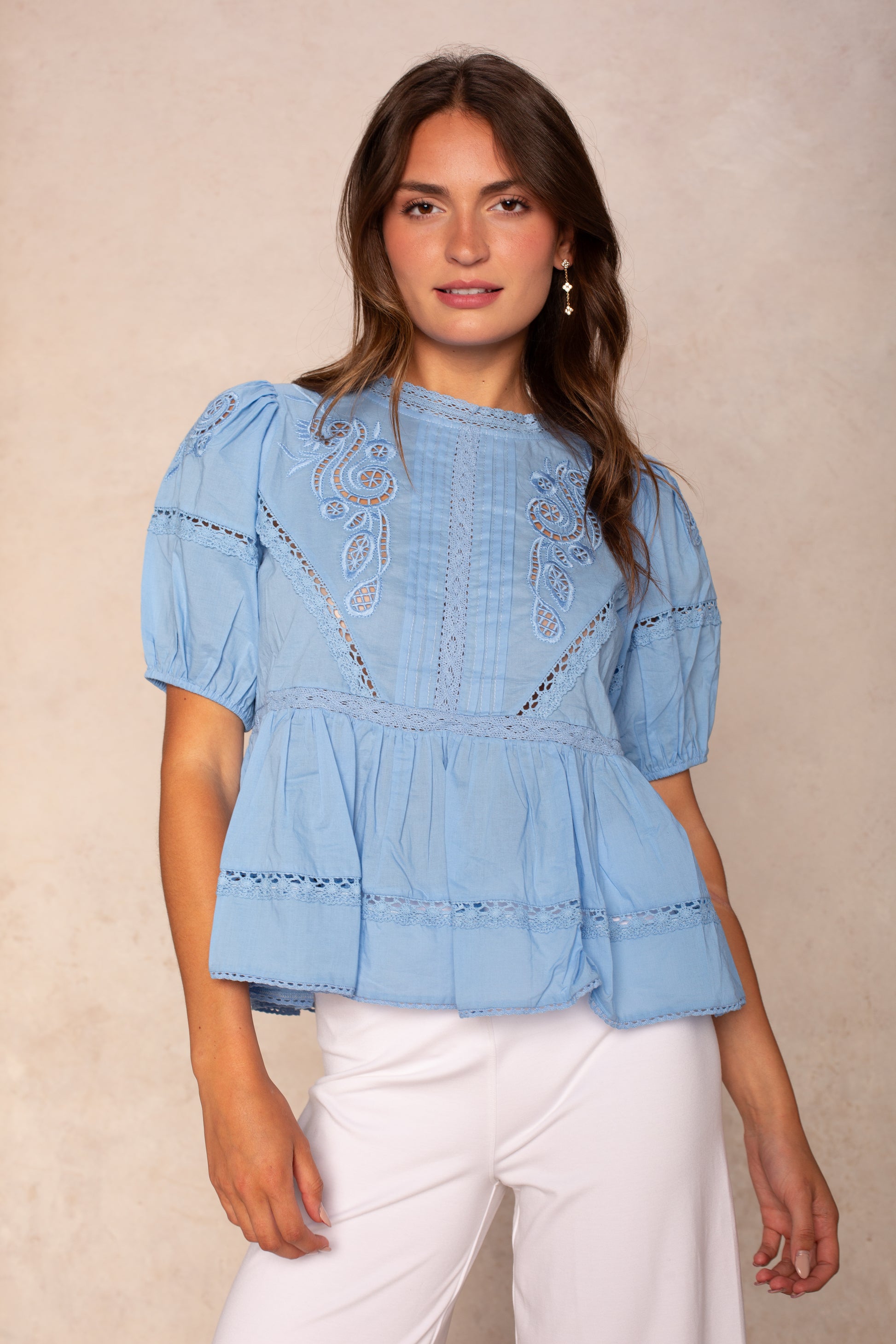 A woman with long brown hair wears the Georgie Rachel Eyelet Short Sleeve Blouse in blue and white pants, standing before a neutral background and smiling slightly.