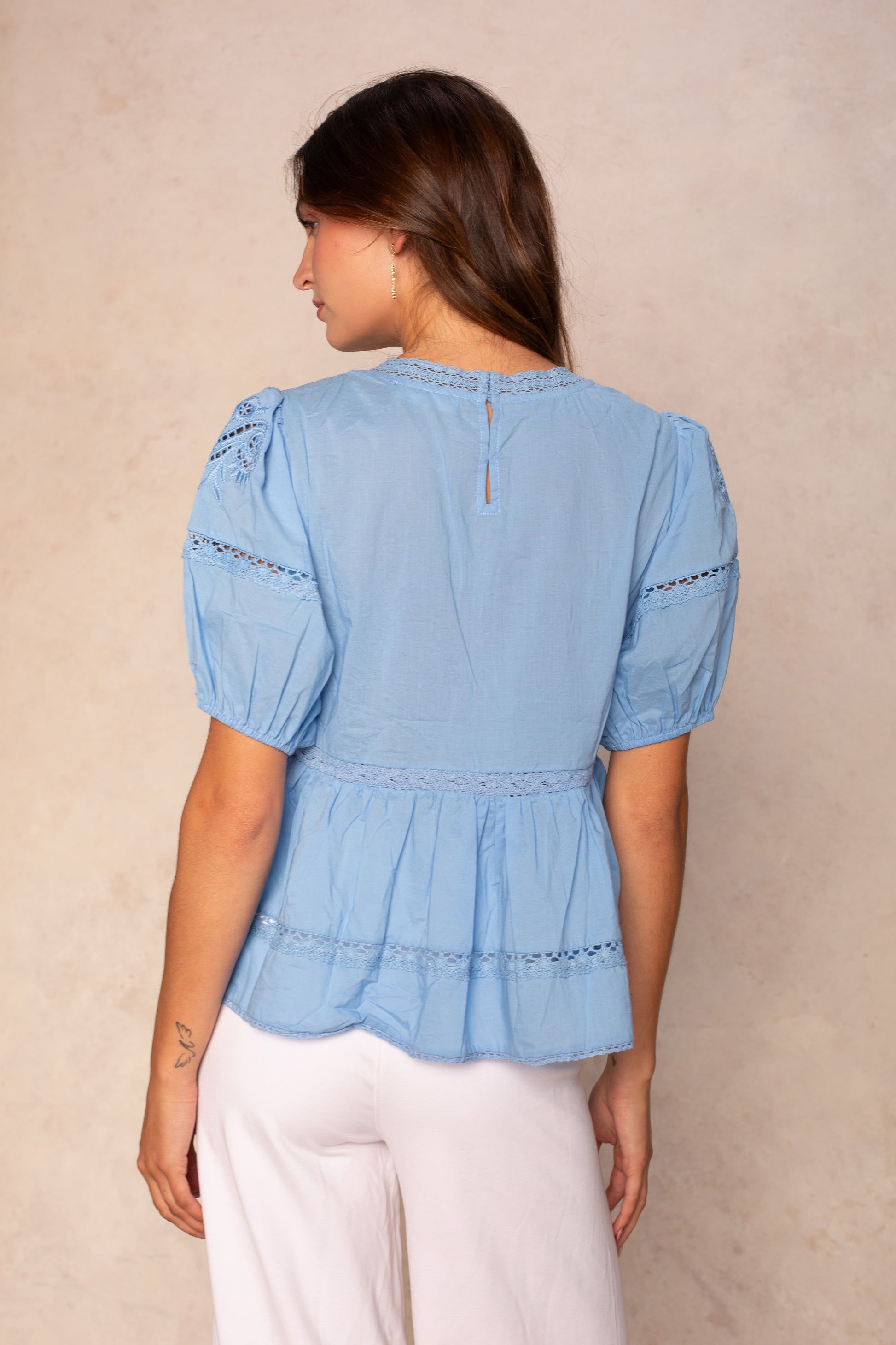 A woman with long brown hair is seen from the back, wearing the Georgie Rachel Eyelet Short Sleeve Blouse in light blue and white pants, standing against a beige background.