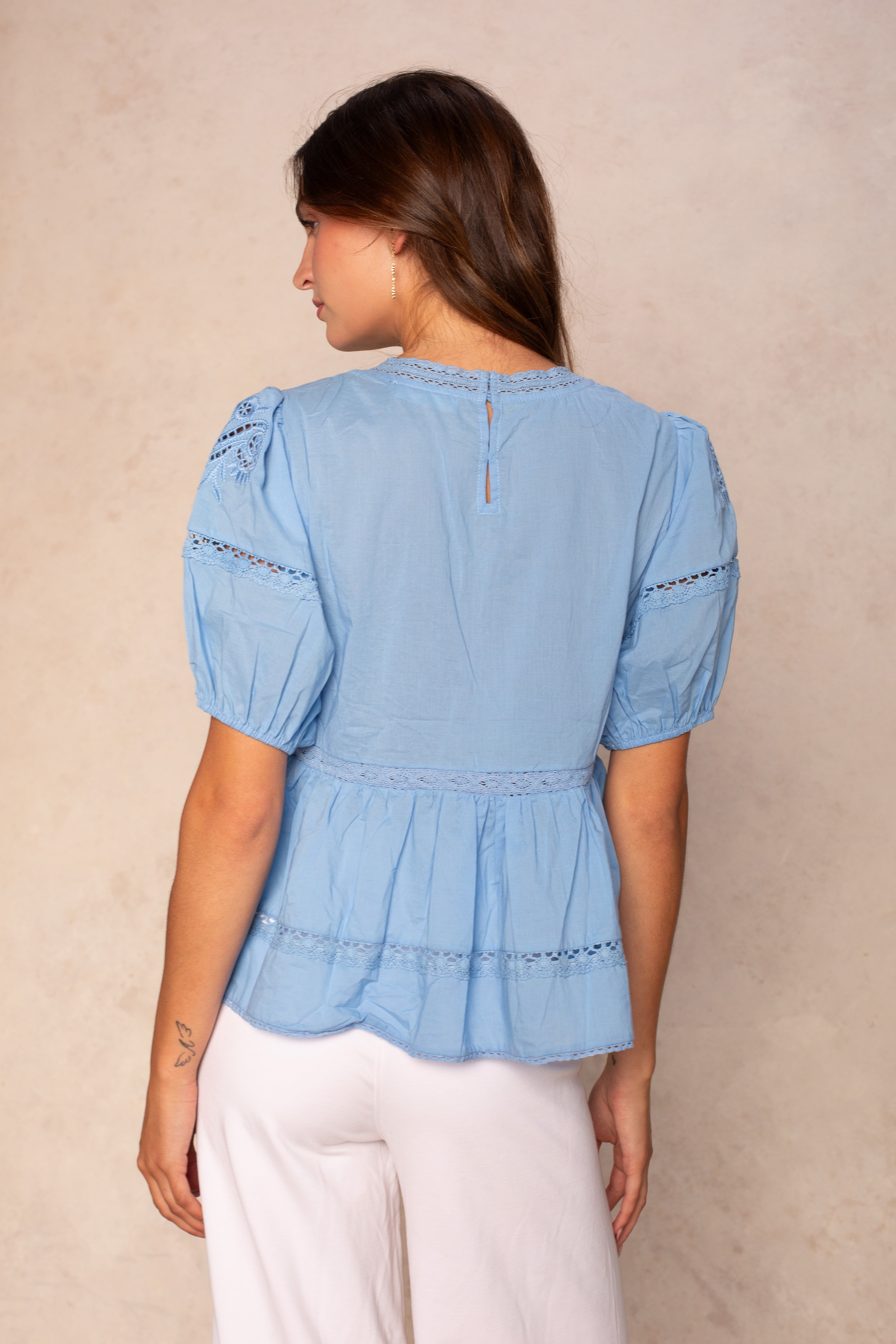 A woman with long brown hair is seen from the back, wearing the Georgie Rachel Eyelet Short Sleeve Blouse in light blue and white pants, standing against a beige background.