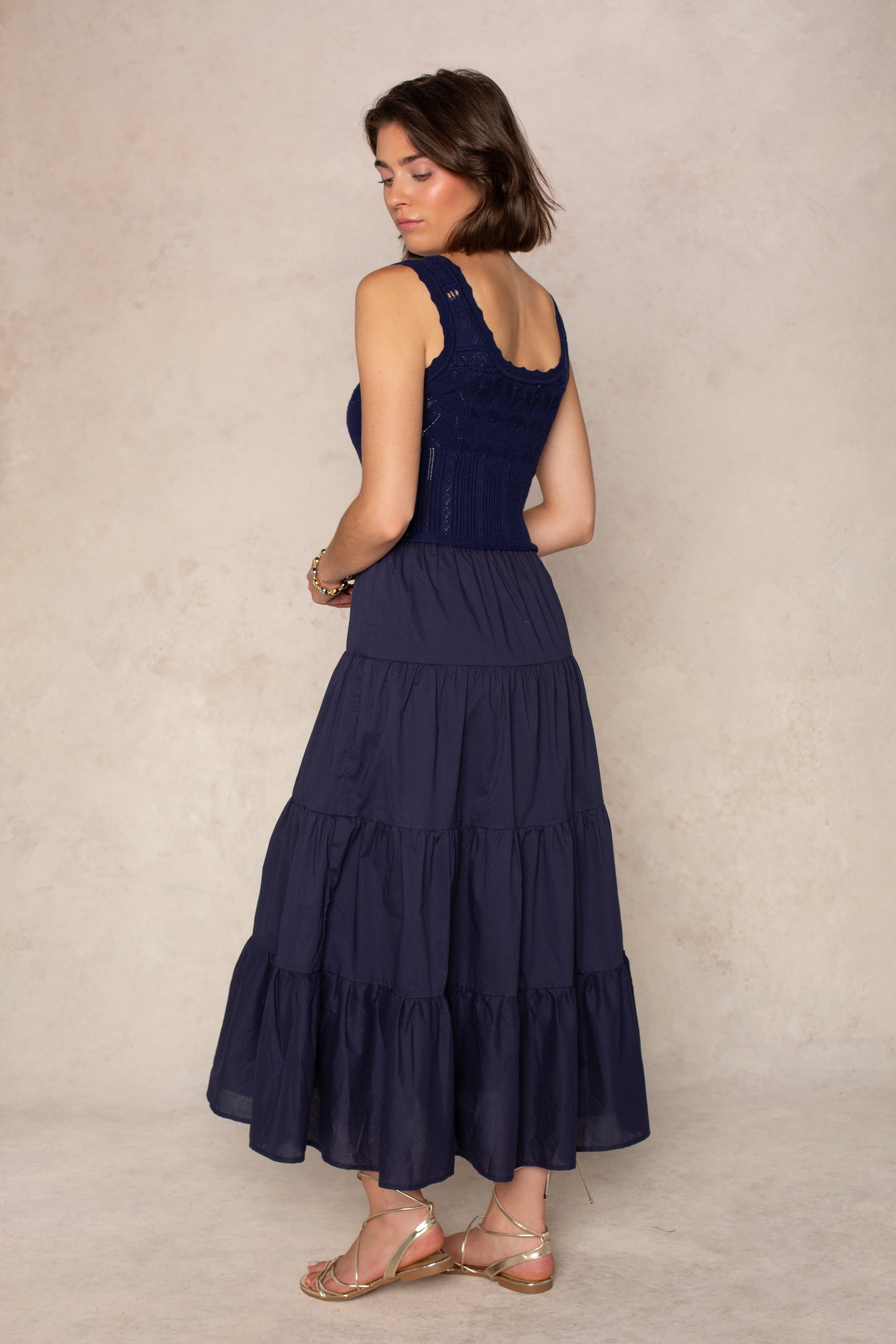 A woman stands sideways and looks down, wearing the Georgie Aria Midi Dress—a navy dress featuring a crochet bodice and tiered skirt—paired with gold sandals, in front of a beige textured background.