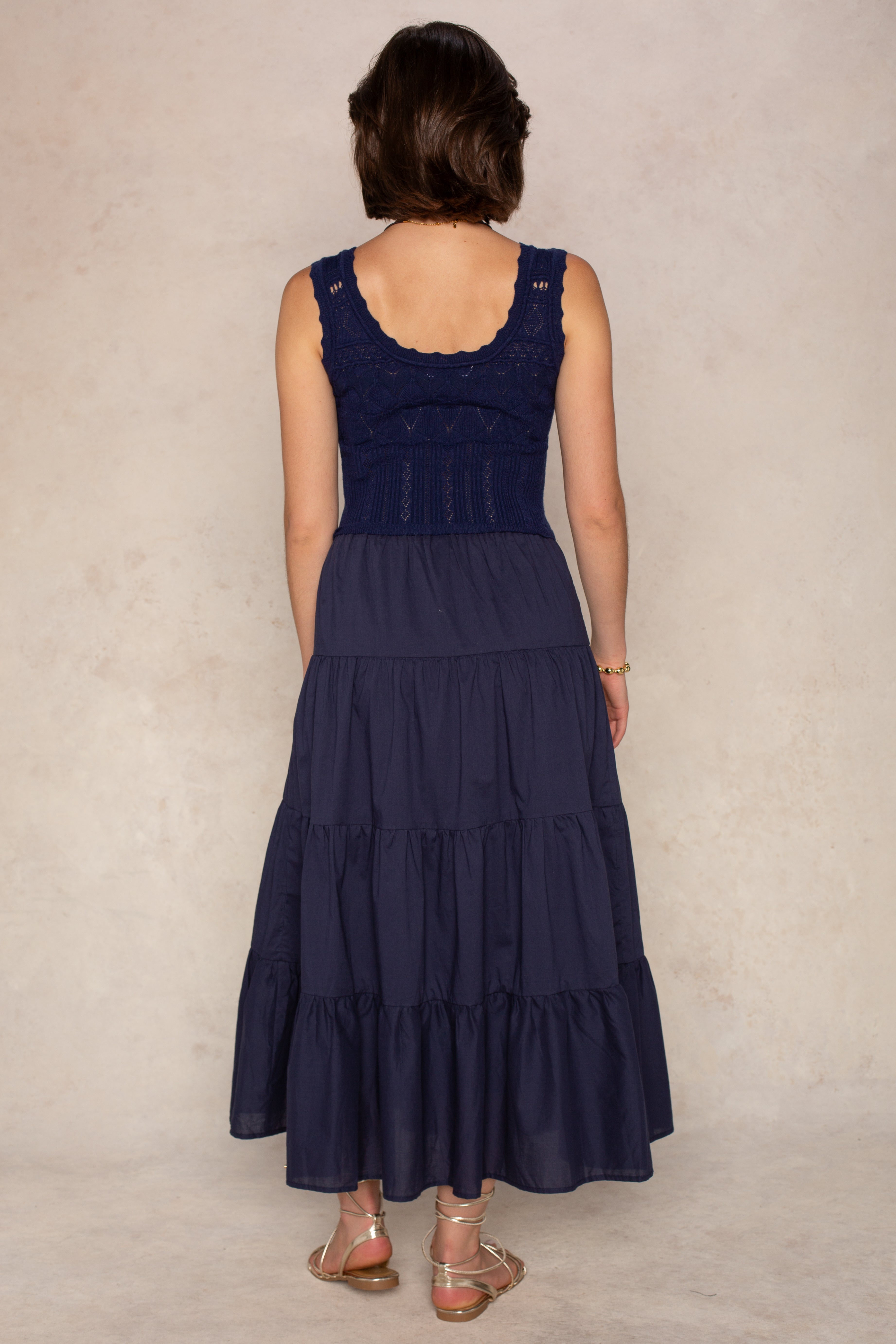 A woman with short brown hair stands facing away, wearing the Georgie Aria Midi Dress—a navy dress with a tiered skirt and delicate crochet bodice. She pairs it with flat sandals against a light beige background.