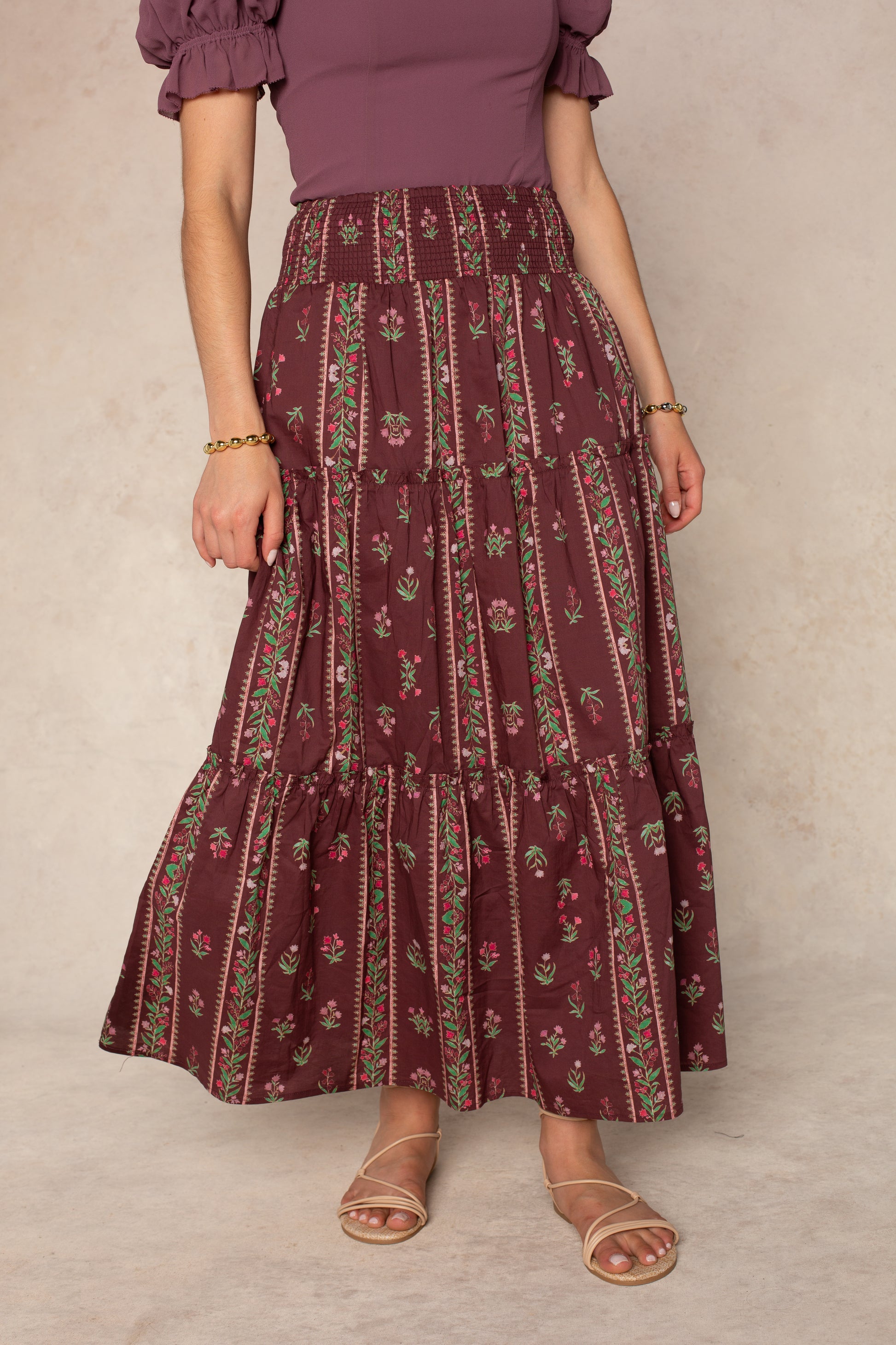 A person wearing the Georgie Melisse Maxi Skirt in burgundy—a bohemian-style maxi skirt with a smocked waistband and floral stripe print—paired with a mauve short-sleeve top, tan sandals, and gold bracelets stands against a neutral background.