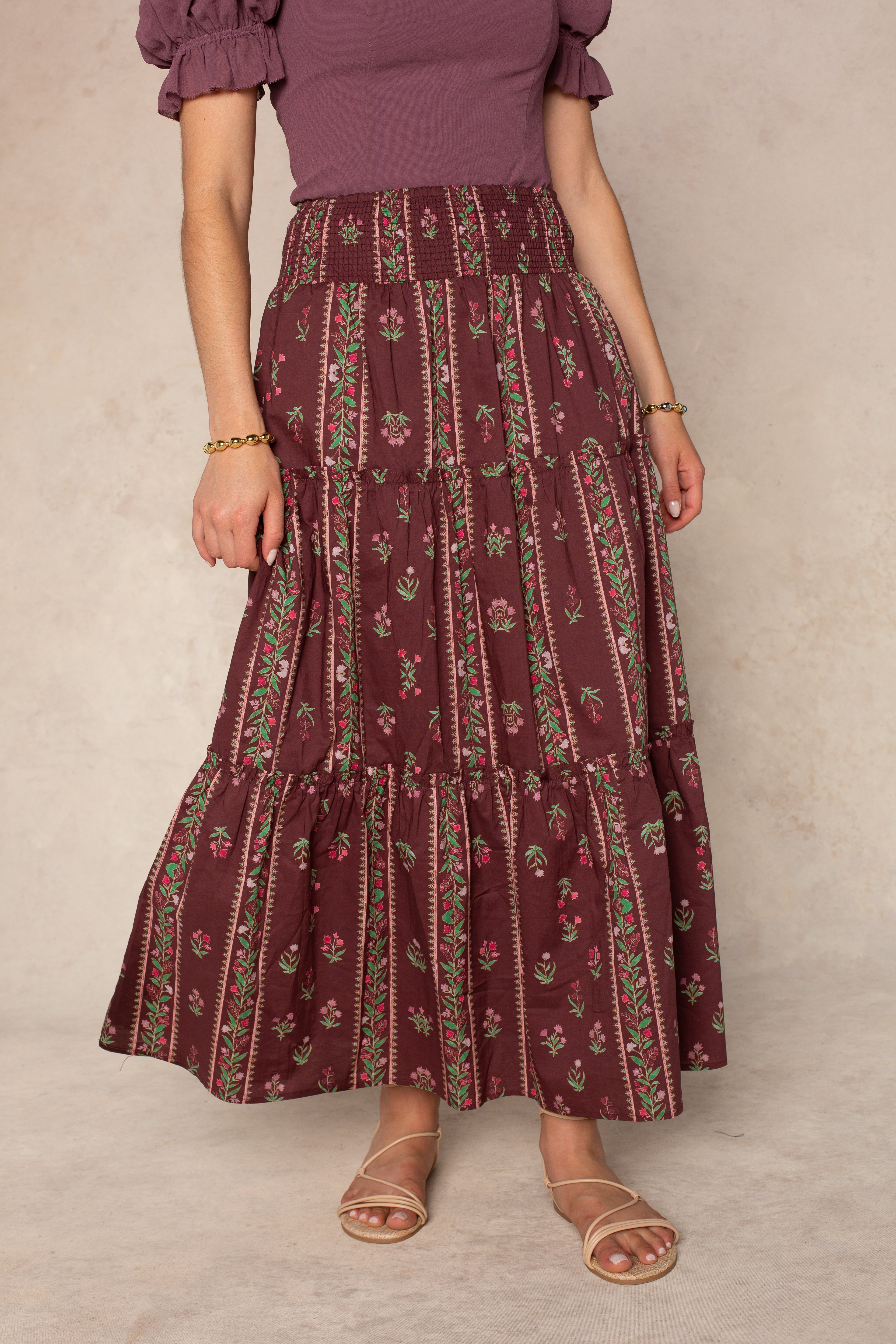 A person wearing the Georgie Melisse Maxi Skirt in burgundy—a bohemian-style maxi skirt with a smocked waistband and floral stripe print—paired with a mauve short-sleeve top, tan sandals, and gold bracelets stands against a neutral background.
