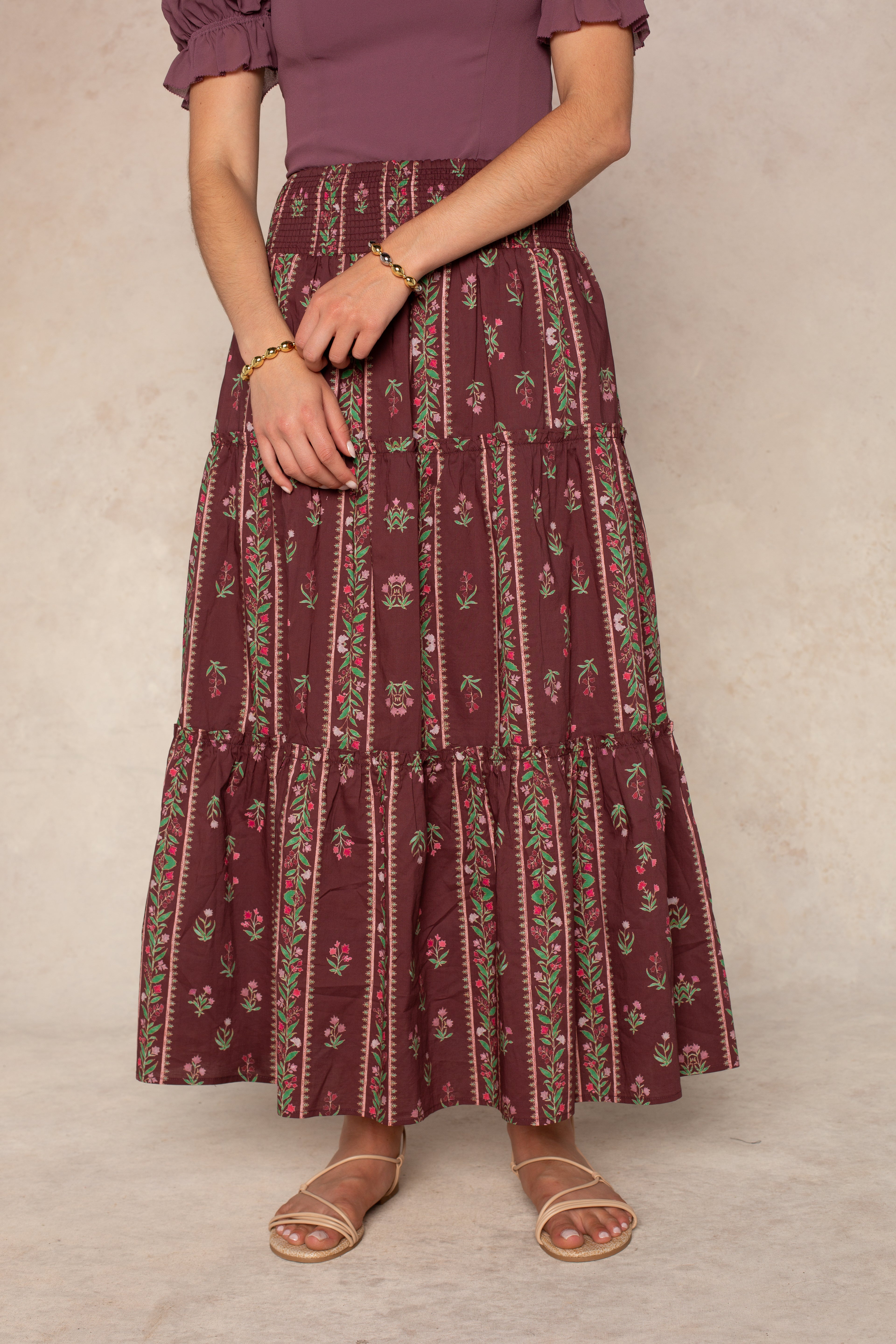 A person wearing the Georgie Melisse Maxi Skirt, featuring a maroon floral stripe print, tiered silhouette, and smocked waistband, paired with the matching short-sleeve top, gold bracelet, and nude strappy sandals stands against a neutral backdrop.