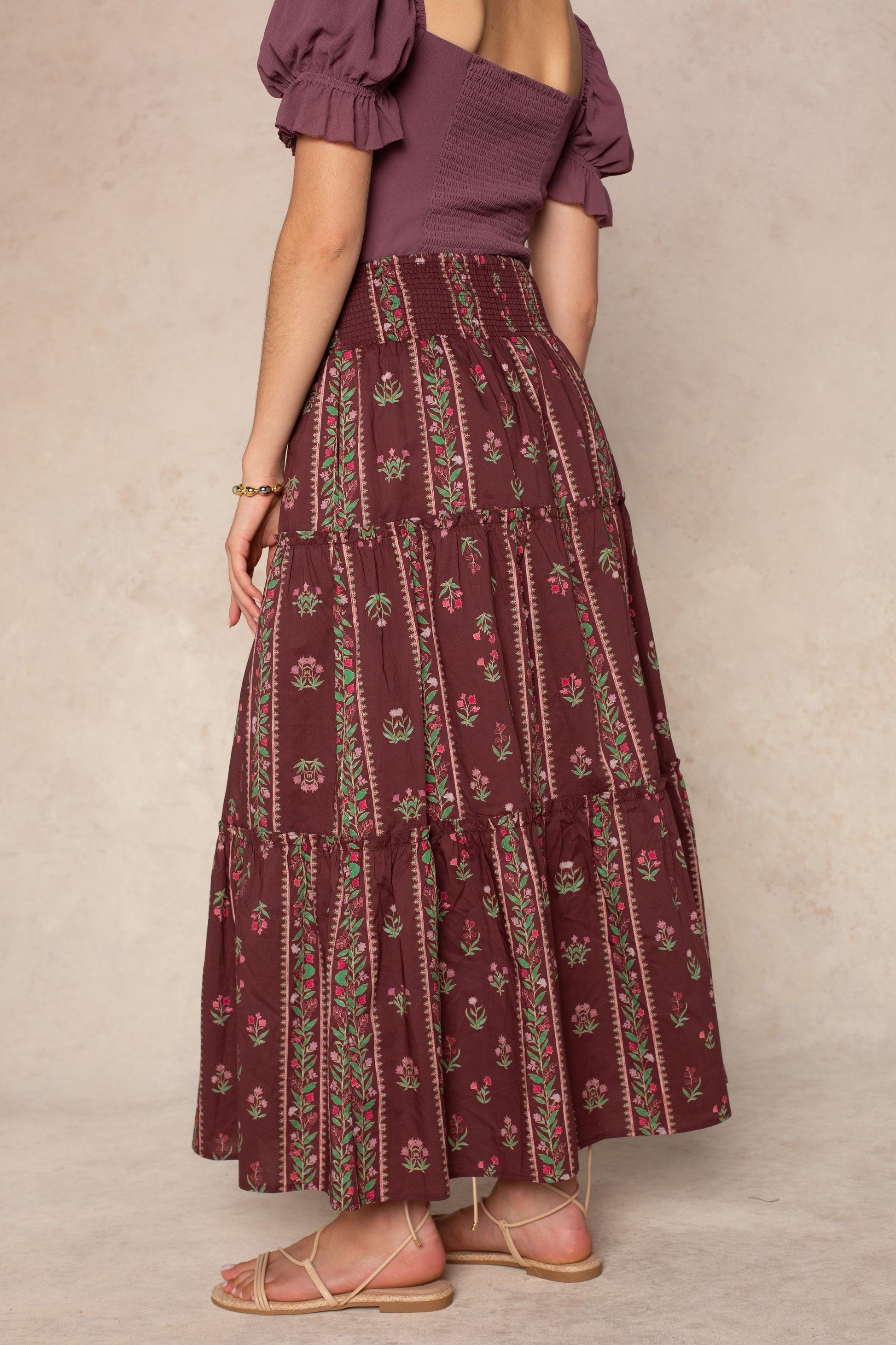 A woman stands barefoot in sandals, facing away from the camera against a beige background, wearing a mauve puff-sleeve top paired with the Georgie Melisse Maxi Skirt featuring a bohemian floral stripe print.