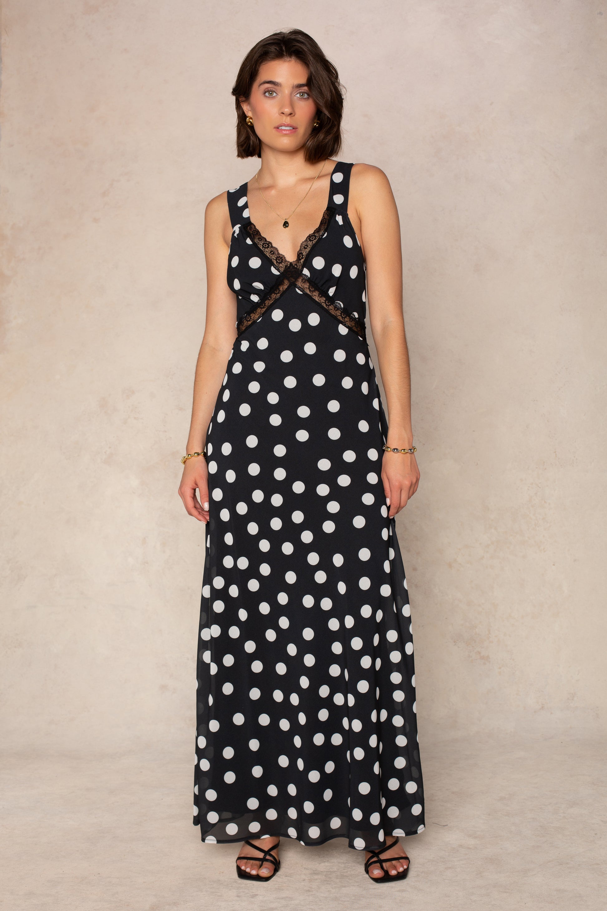 A woman with short brown hair stands against a beige background, wearing the Georgie Mia Lace Slip Maxi Dress in BLACK POLKA DOT paired with black sandals and gold jewelry.