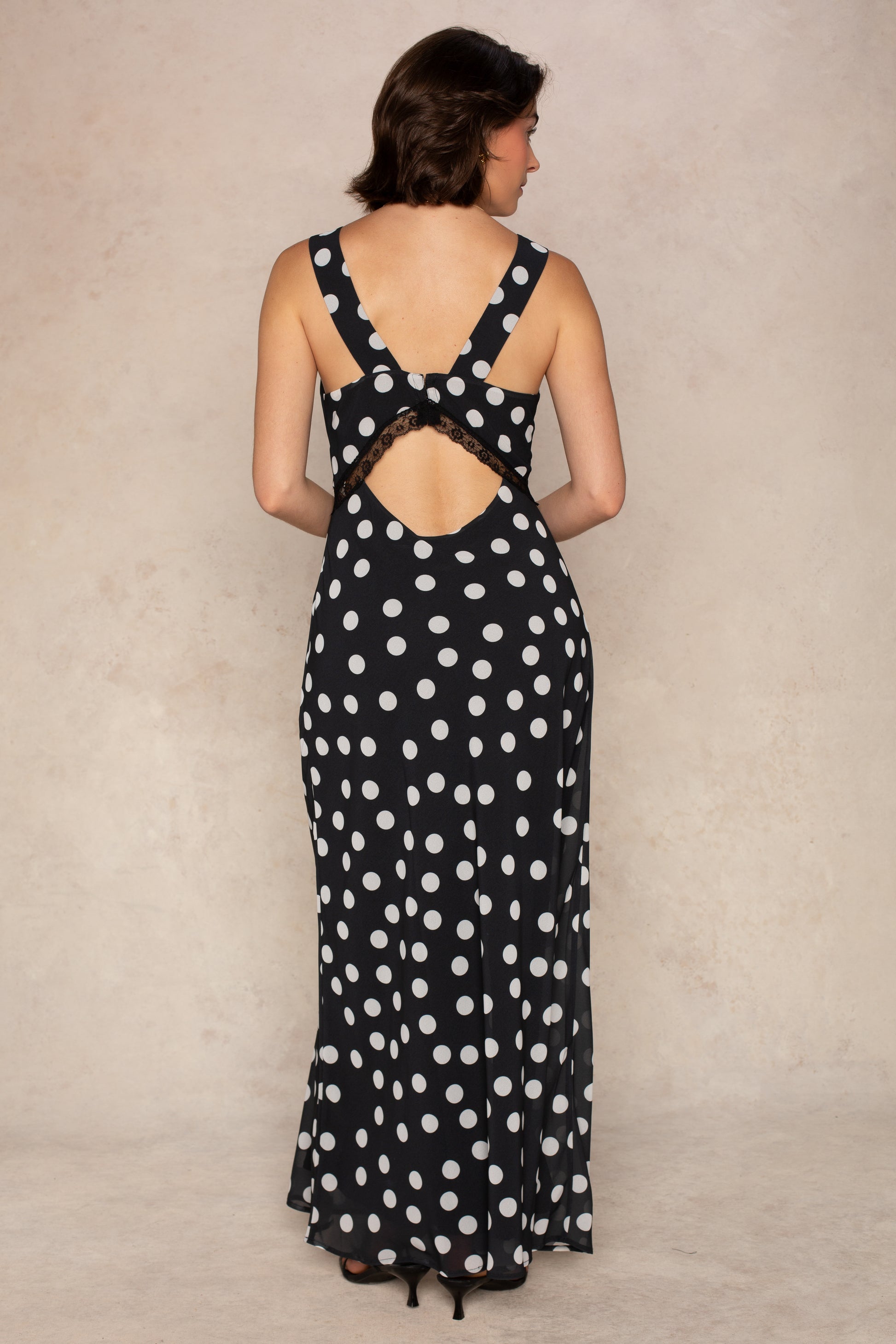 A woman stands facing away in the Mia Lace Slip Maxi Dress by Georgie—a black, sleeveless maxi dress adorned with white polka dots, a back cutout, and delicate lace detailing. Perfectly paired with black heels against a light-colored backdrop, this timeless piece is an elegant choice for summer.