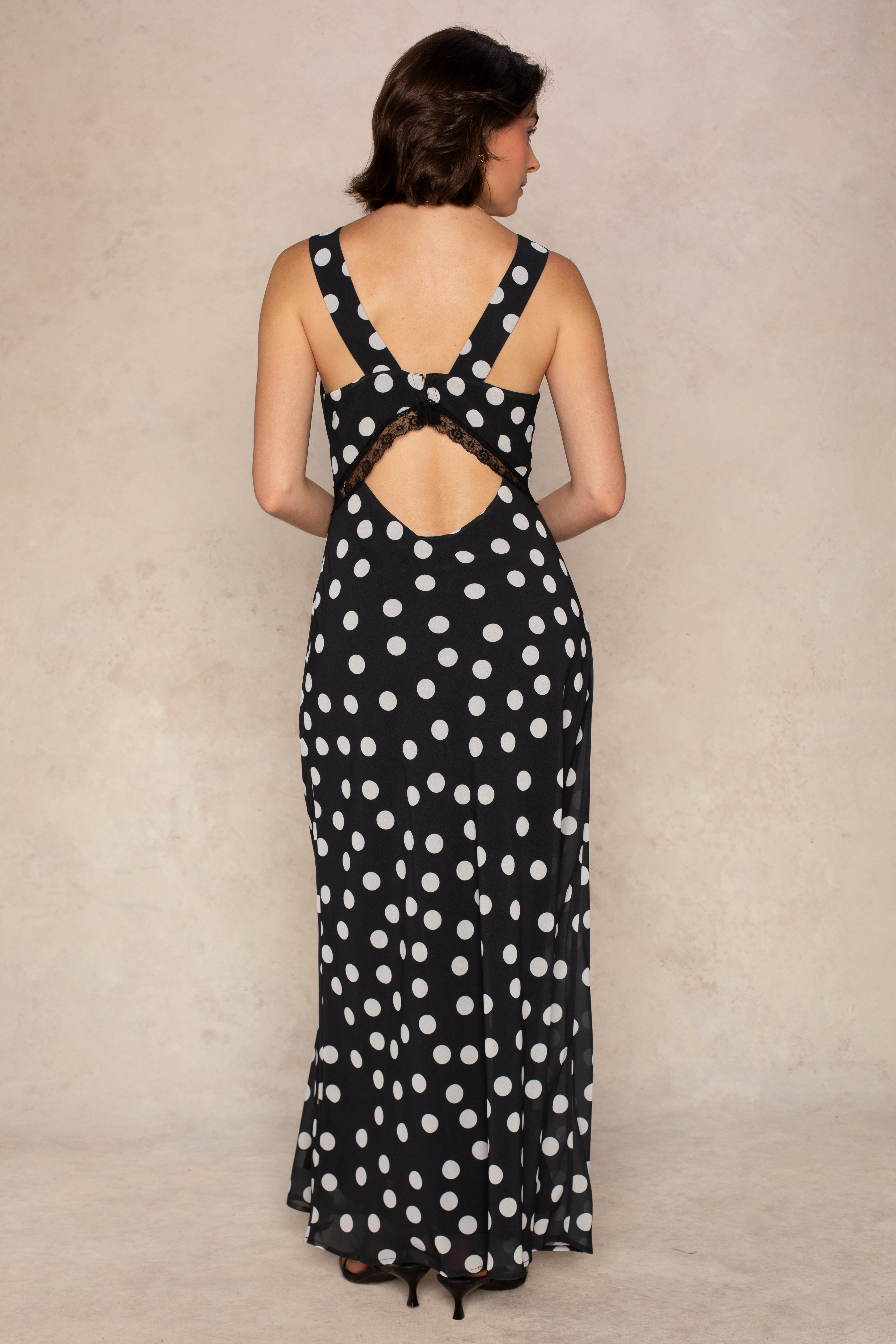 A woman stands facing away in the Mia Lace Slip Maxi Dress by Georgie—a black, sleeveless maxi dress adorned with white polka dots, a back cutout, and delicate lace detailing. Perfectly paired with black heels against a light-colored backdrop, this timeless piece is an elegant choice for summer.