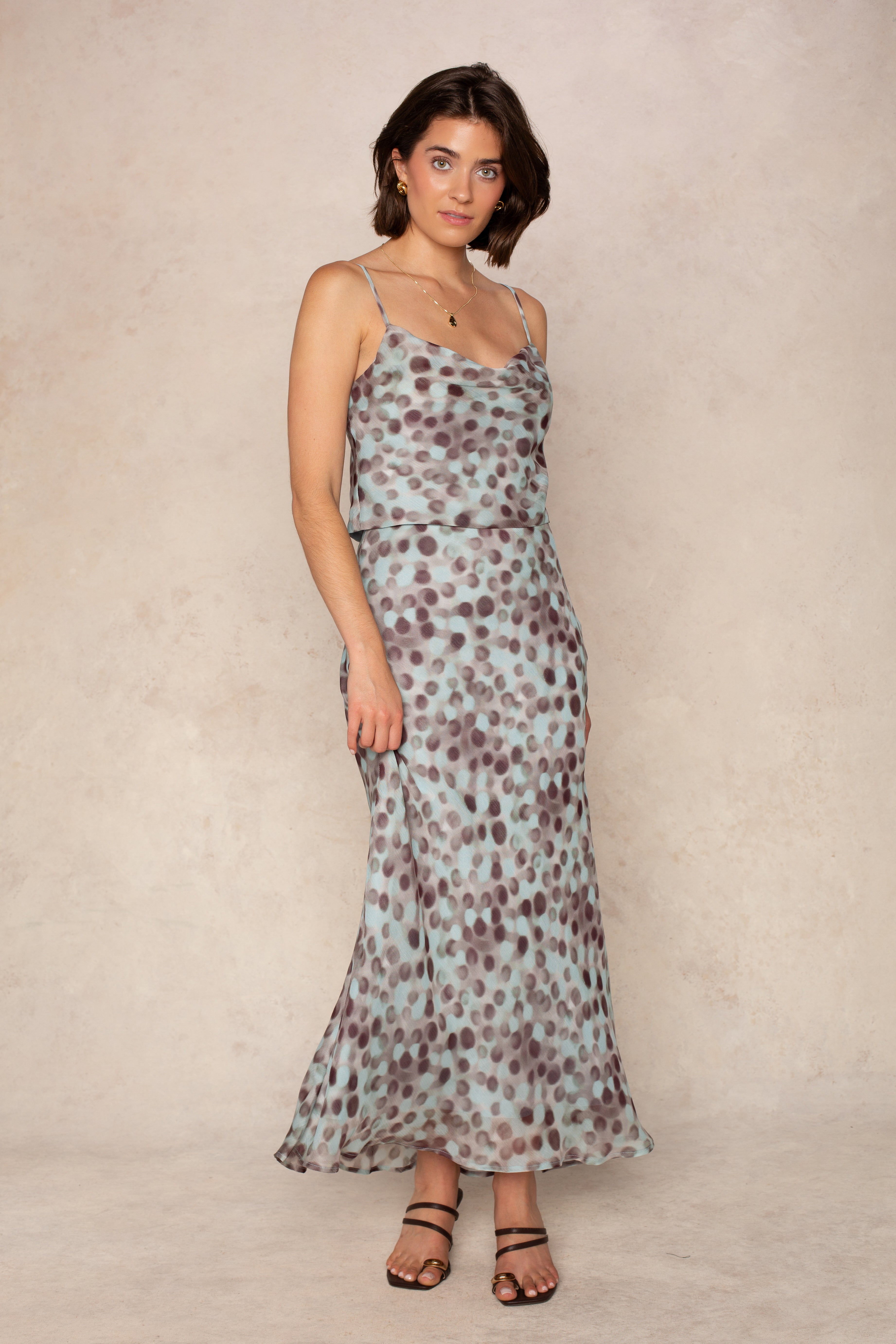 A woman stands against a beige textured background wearing the Isla Naomi Chiffon Cami, a light blue dress with dark polka dots, adjustable spaghetti straps, and a flowy skirt. She pairs it with black strappy sandals.