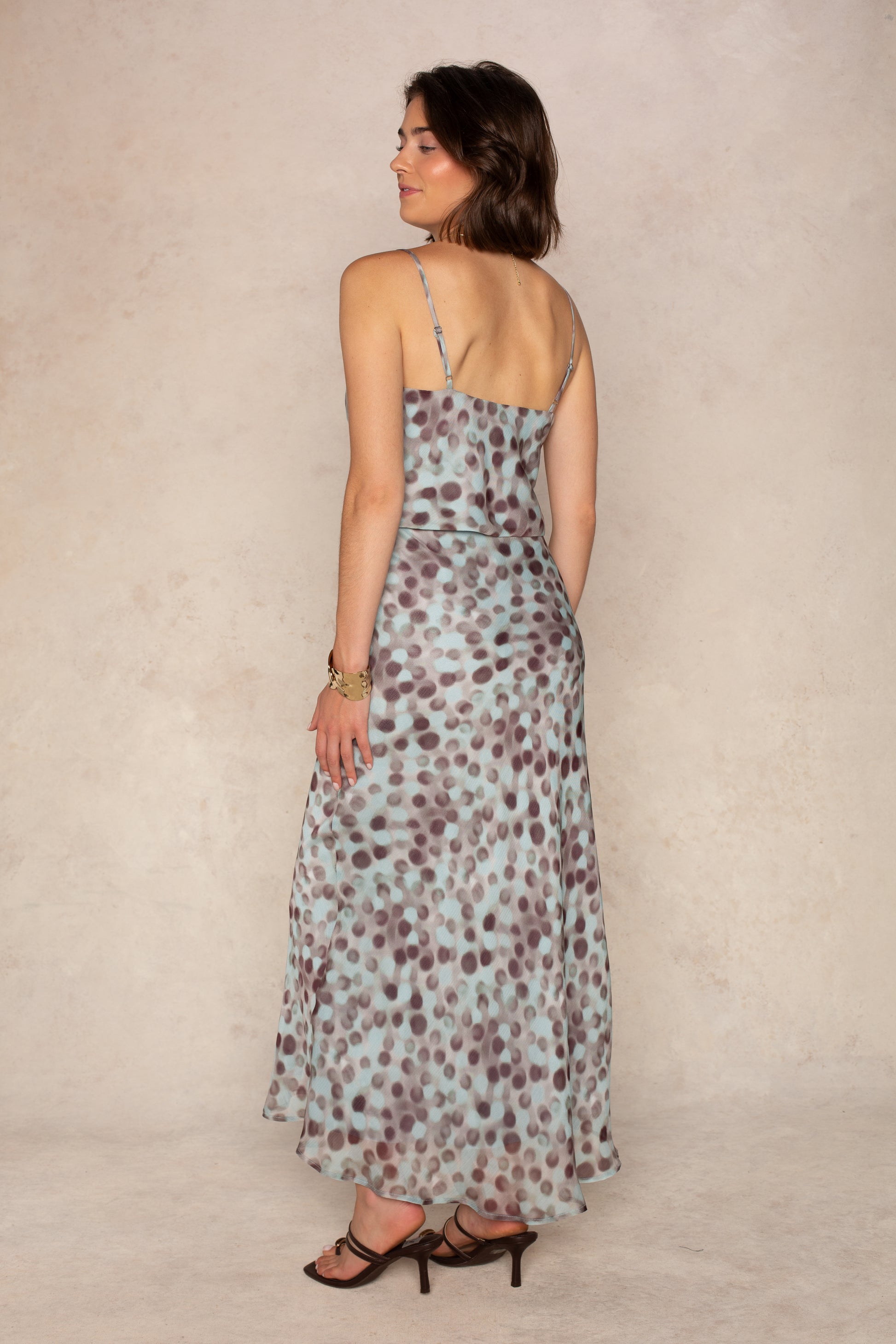 A woman stands sideways, glancing back over her shoulder, wearing the Isla Naomi Chiffon Midi Skirt—a sleeveless, light blue piece featuring an abstract dot print—paired with strappy black heels against a neutral textured background.