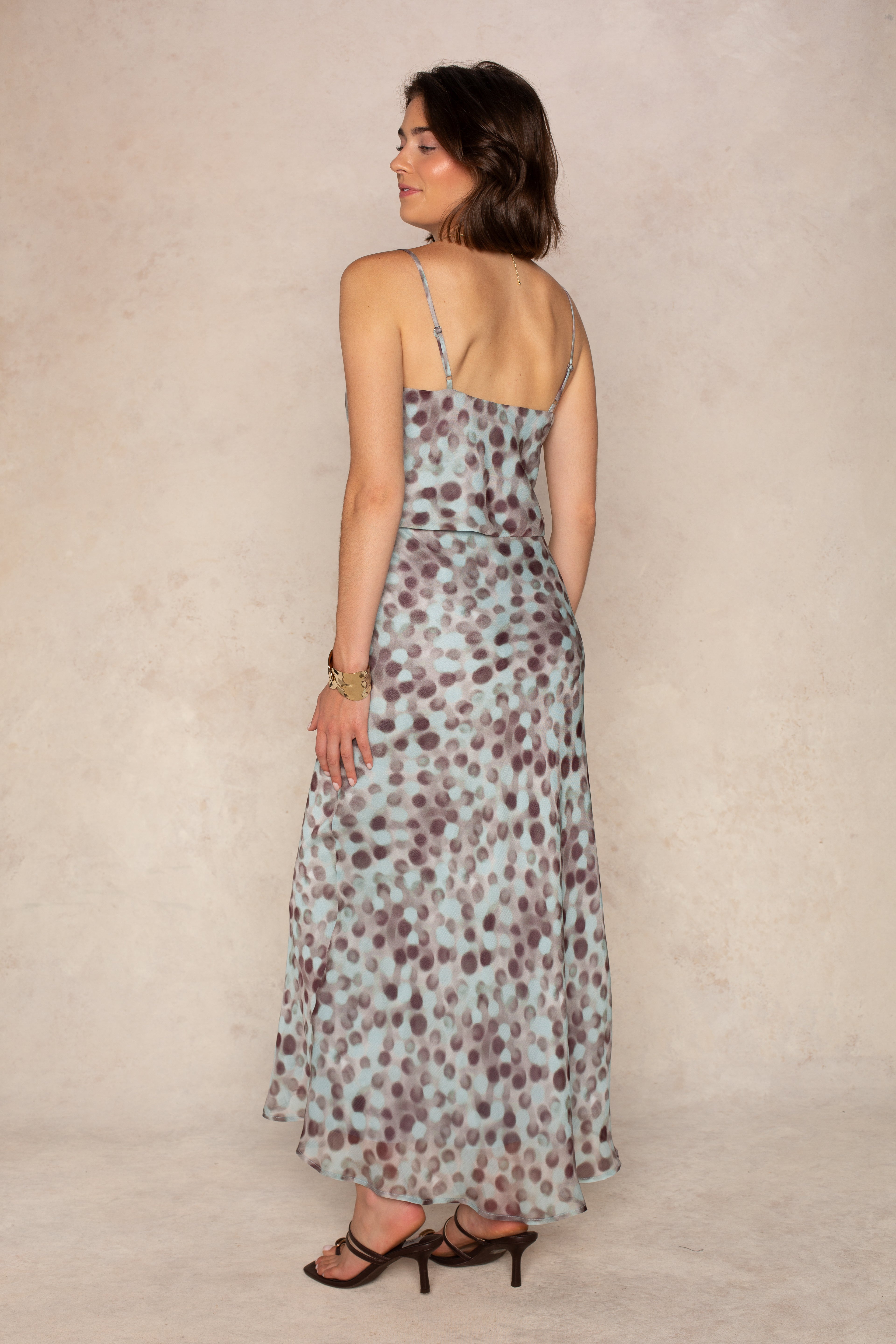 A woman stands sideways, glancing back over her shoulder, wearing the Isla Naomi Chiffon Midi Skirt—a sleeveless, light blue piece featuring an abstract dot print—paired with strappy black heels against a neutral textured background.