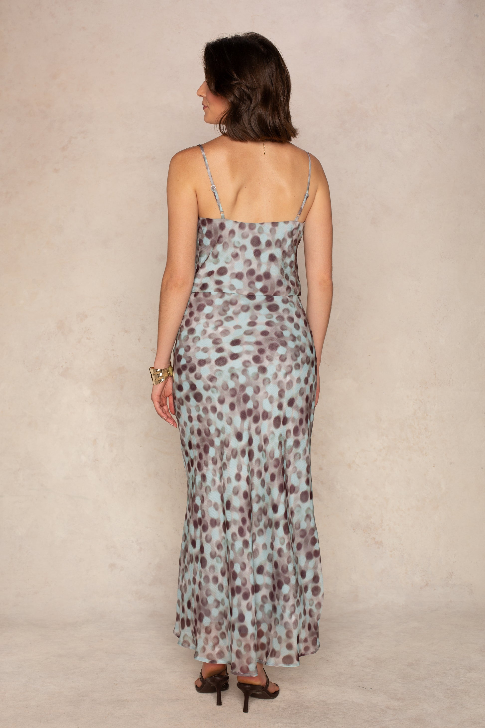 A woman stands on a light floor against a neutral background, facing away from the camera with her hands at her sides, wearing the Isla Naomi Chiffon Cami—an ankle-length, sleeveless dress featuring adjustable spaghetti straps and a blue and brown polka dot pattern.