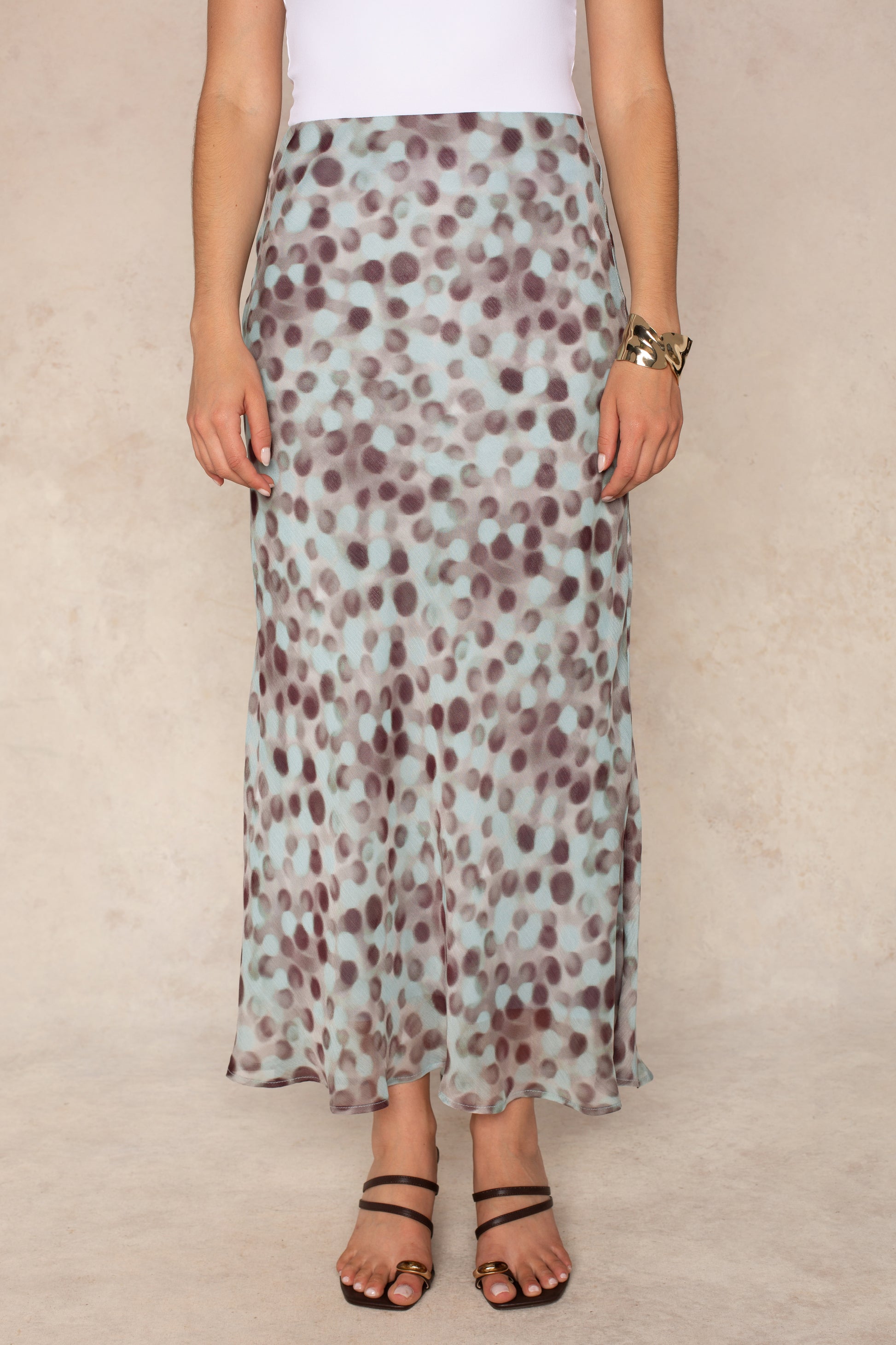 A person wears a white top and the Isla Naomi Chiffon Midi Skirt in Aqua Grey, which features a high-rise waistband and an abstract dot print in light blue and brown tones. The outfit is paired with black sandals and a gold bracelet, set against a neutral background.