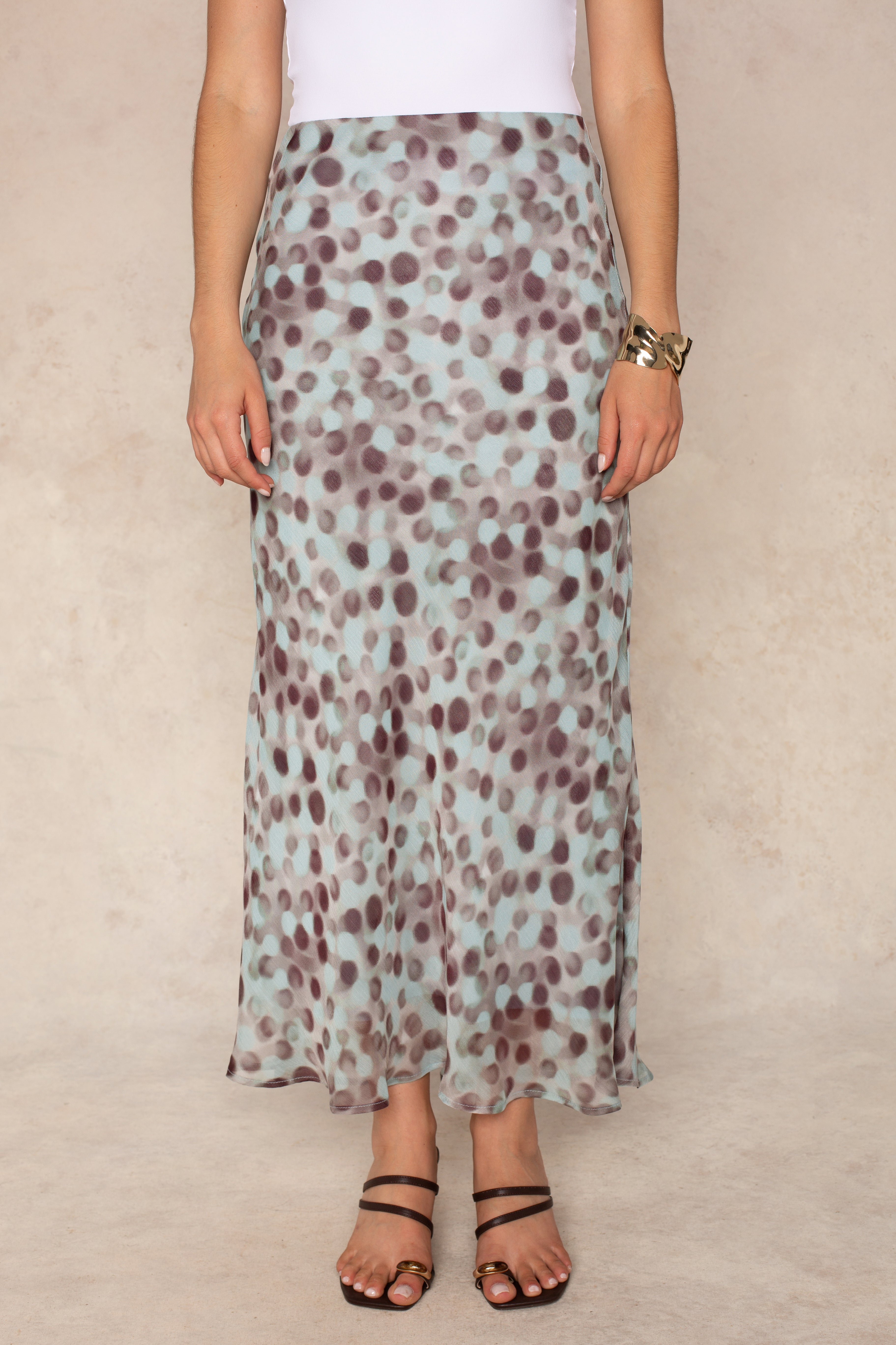 A person wears a white top and the Isla Naomi Chiffon Midi Skirt in Aqua Grey, which features a high-rise waistband and an abstract dot print in light blue and brown tones. The outfit is paired with black sandals and a gold bracelet, set against a neutral background.