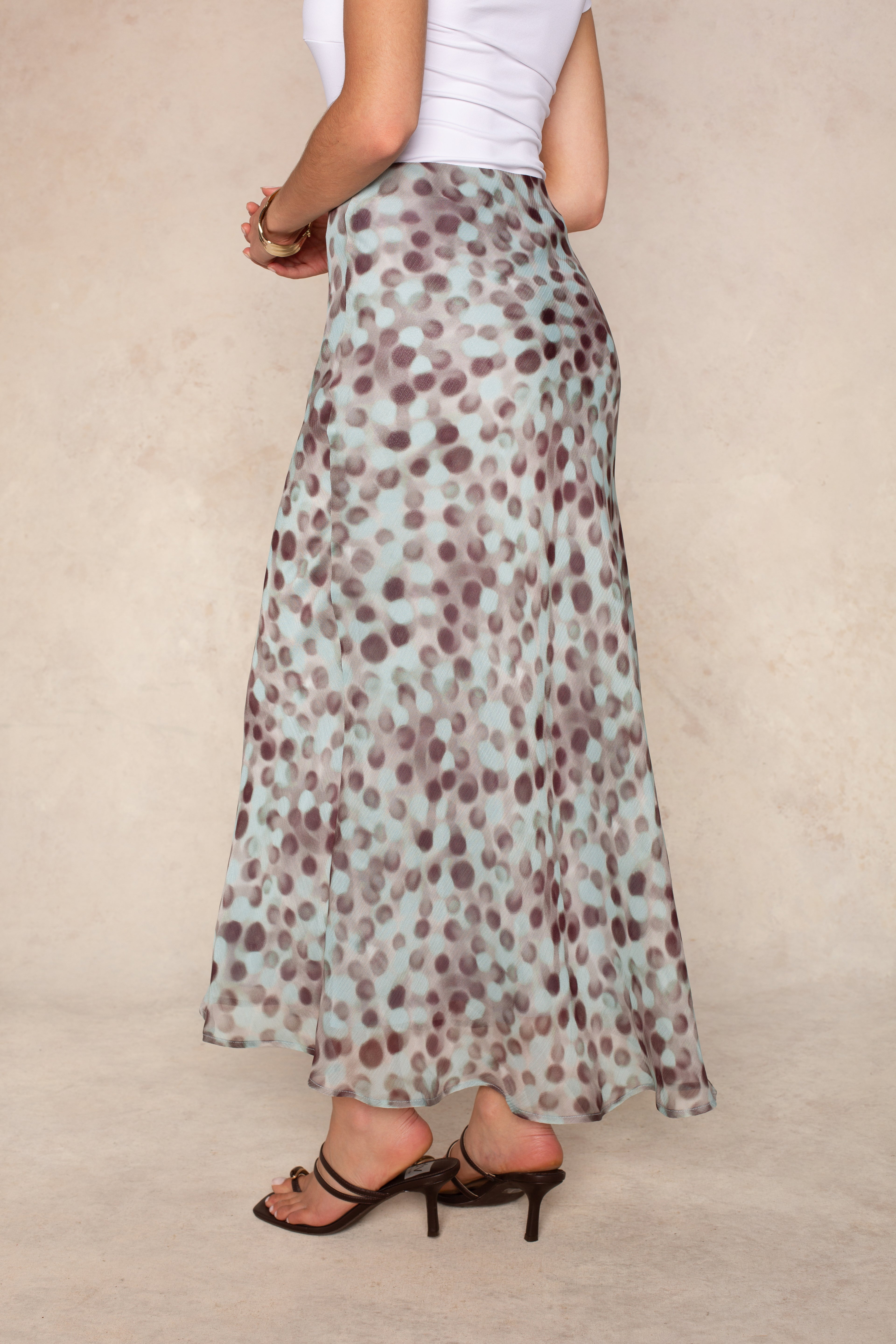 A person wearing a white top and the Naomi Chiffon Midi Skirt by Isla—a long, flowy skirt with an abstract blue and brown dot print and a high-rise waistband—stands in brown heeled sandals against a neutral background.