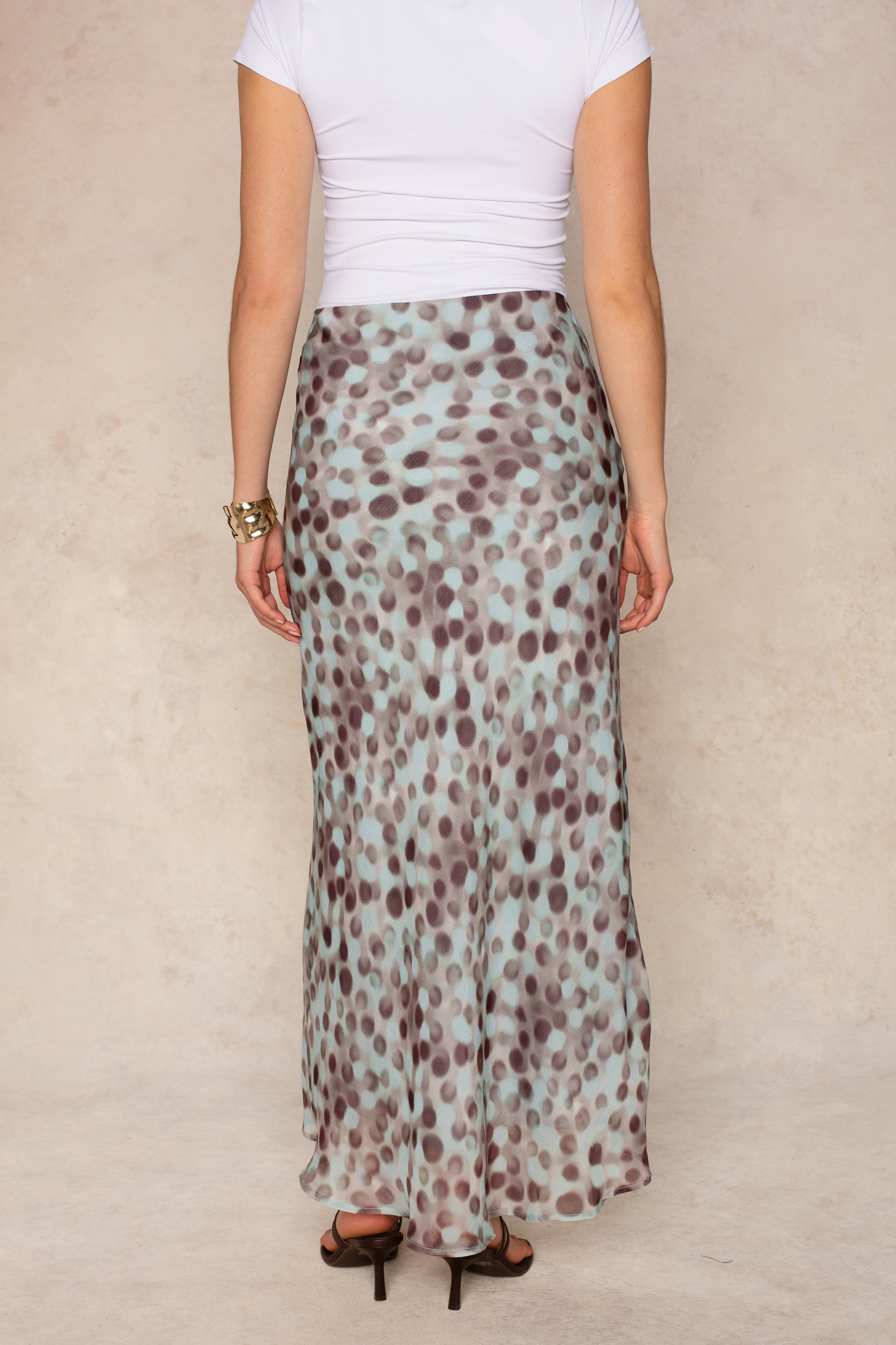 A woman stands with her back to the camera, wearing a fitted white top and the Isla Naomi Chiffon Midi Skirt, which features a blue and brown abstract dot print and a high-rise waistband. She completes the look with black heels and a gold bracelet.