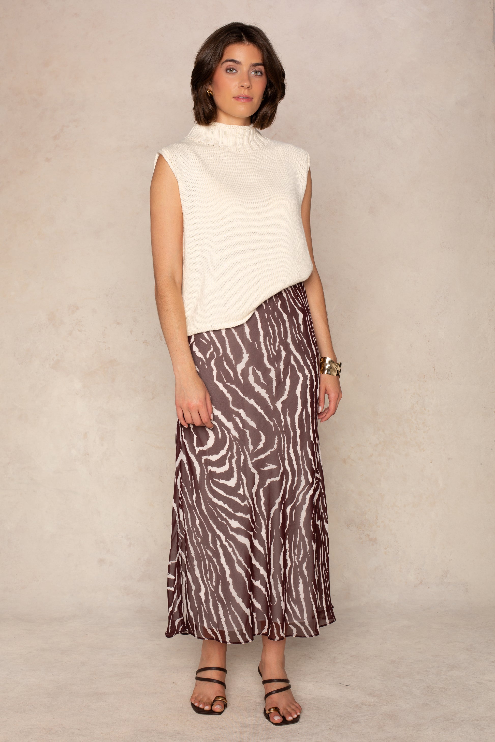 A person stands against a light beige wall wearing a sleeveless ivory turtleneck sweater, the Isla Zenia Chiffon Midi Skirt in BROWN MULTI with a brown and white zebra print and bias-cut silhouette that falls to midi length, paired with black sandals and gold bracelets. The pose is relaxed, set against a neutral background.