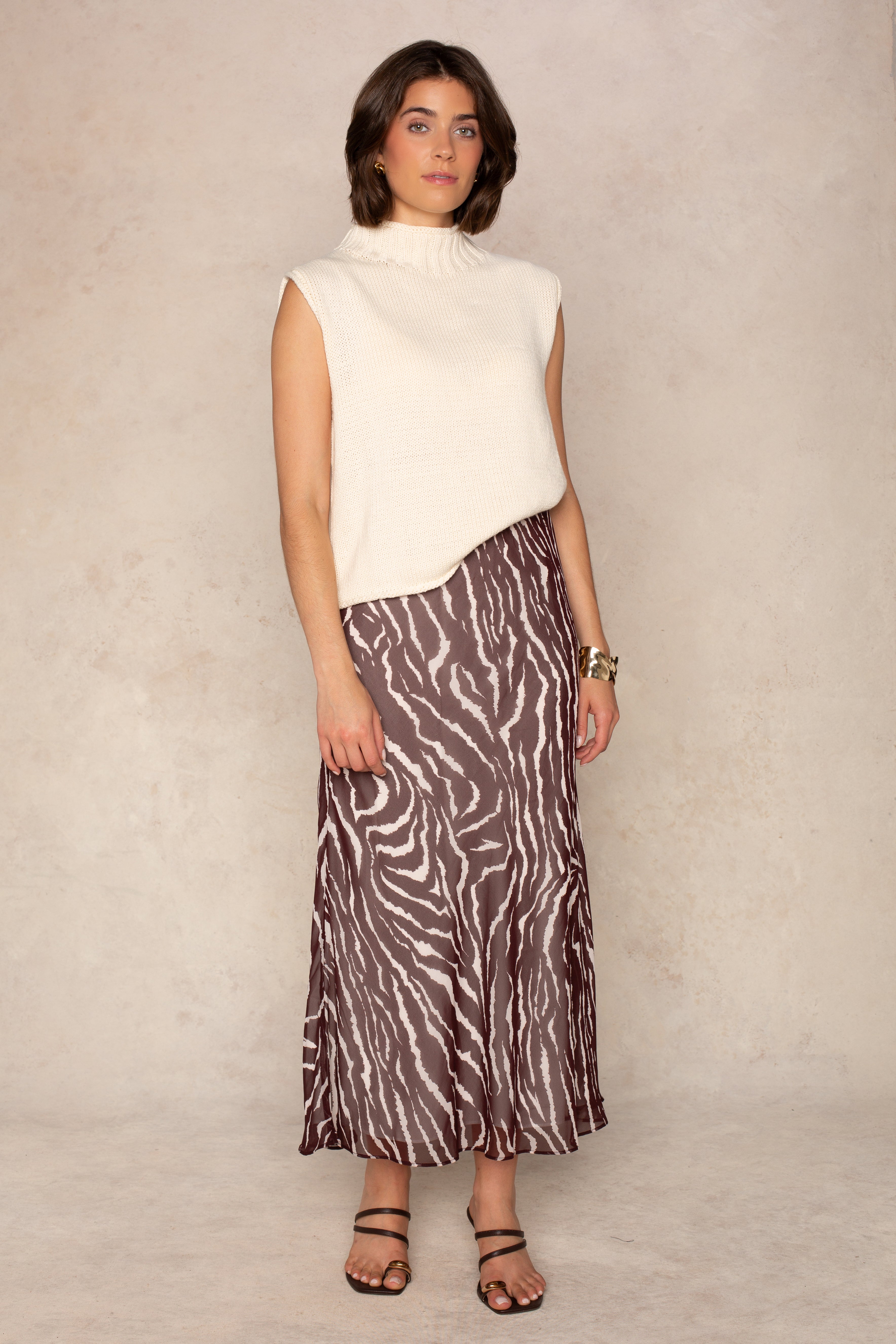 A person stands against a light beige wall wearing a sleeveless ivory turtleneck sweater, the Isla Zenia Chiffon Midi Skirt in BROWN MULTI with a brown and white zebra print and bias-cut silhouette that falls to midi length, paired with black sandals and gold bracelets. The pose is relaxed, set against a neutral background.