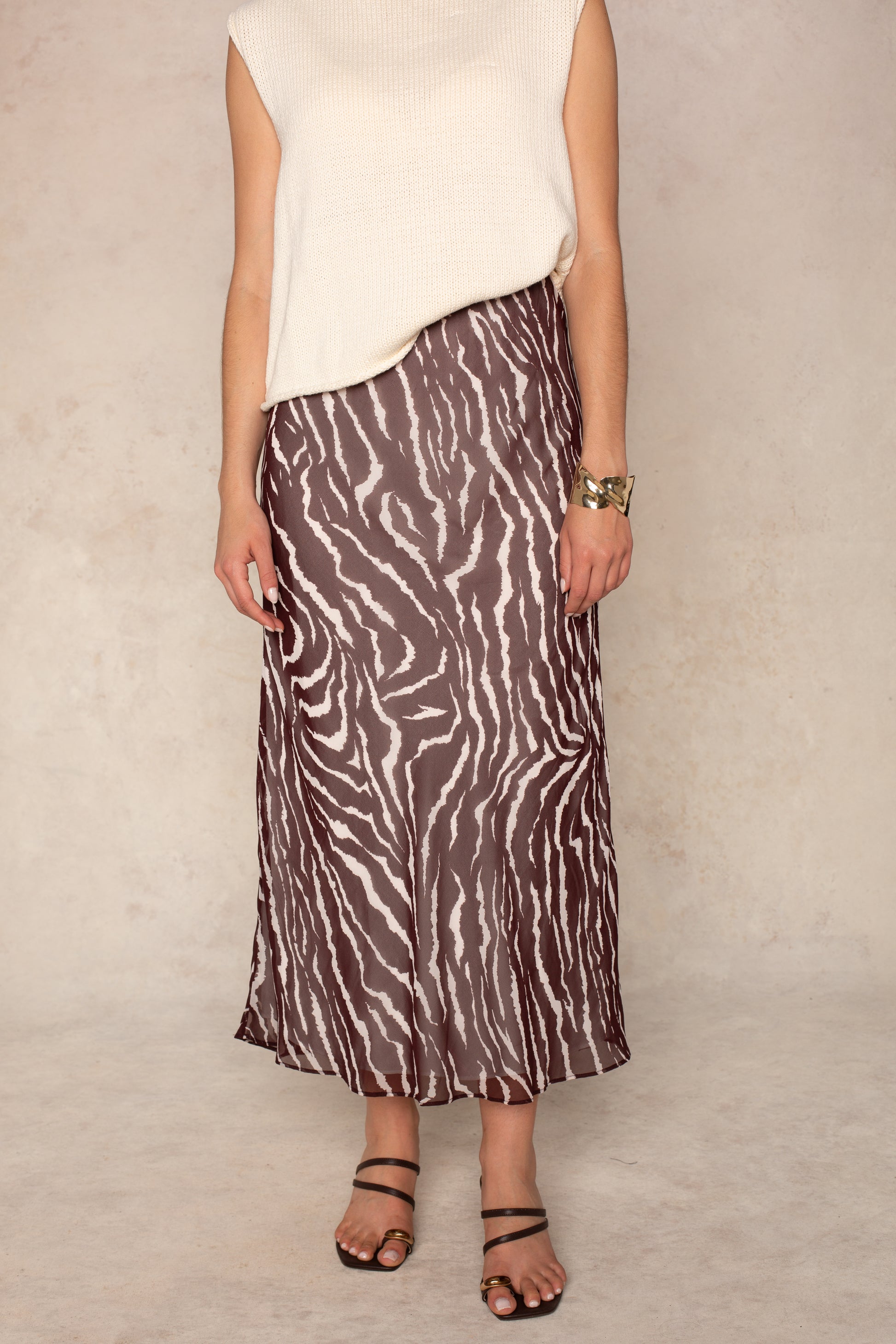 A person models the Isla Zenia Chiffon Midi Skirt, featuring a brown and white zebra print and a flattering bias-cut silhouette, styled with a sleeveless cream top, strappy sandals, and a bracelet against a light background. Their face is not visible.