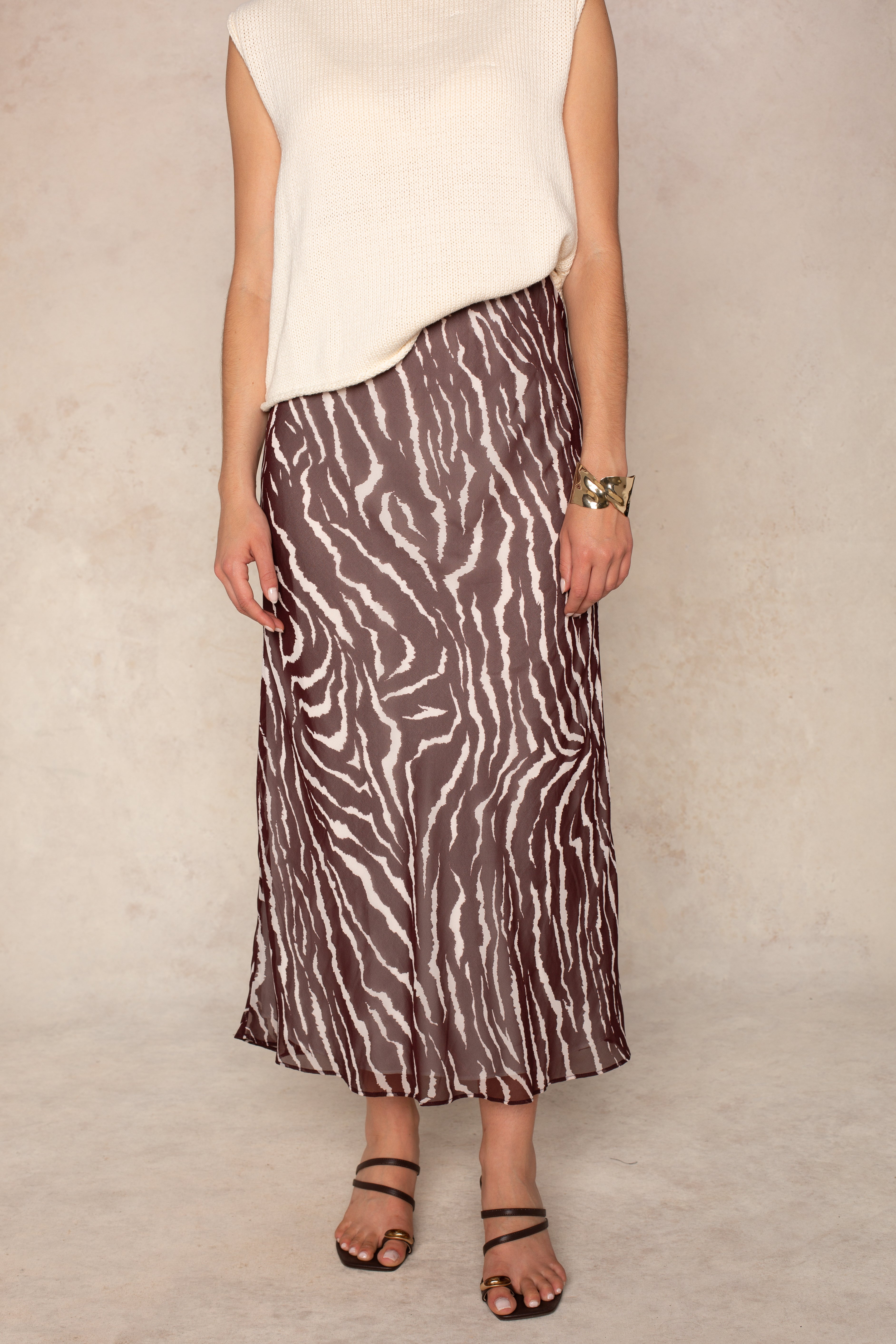A person models the Isla Zenia Chiffon Midi Skirt, featuring a brown and white zebra print and a flattering bias-cut silhouette, styled with a sleeveless cream top, strappy sandals, and a bracelet against a light background. Their face is not visible.