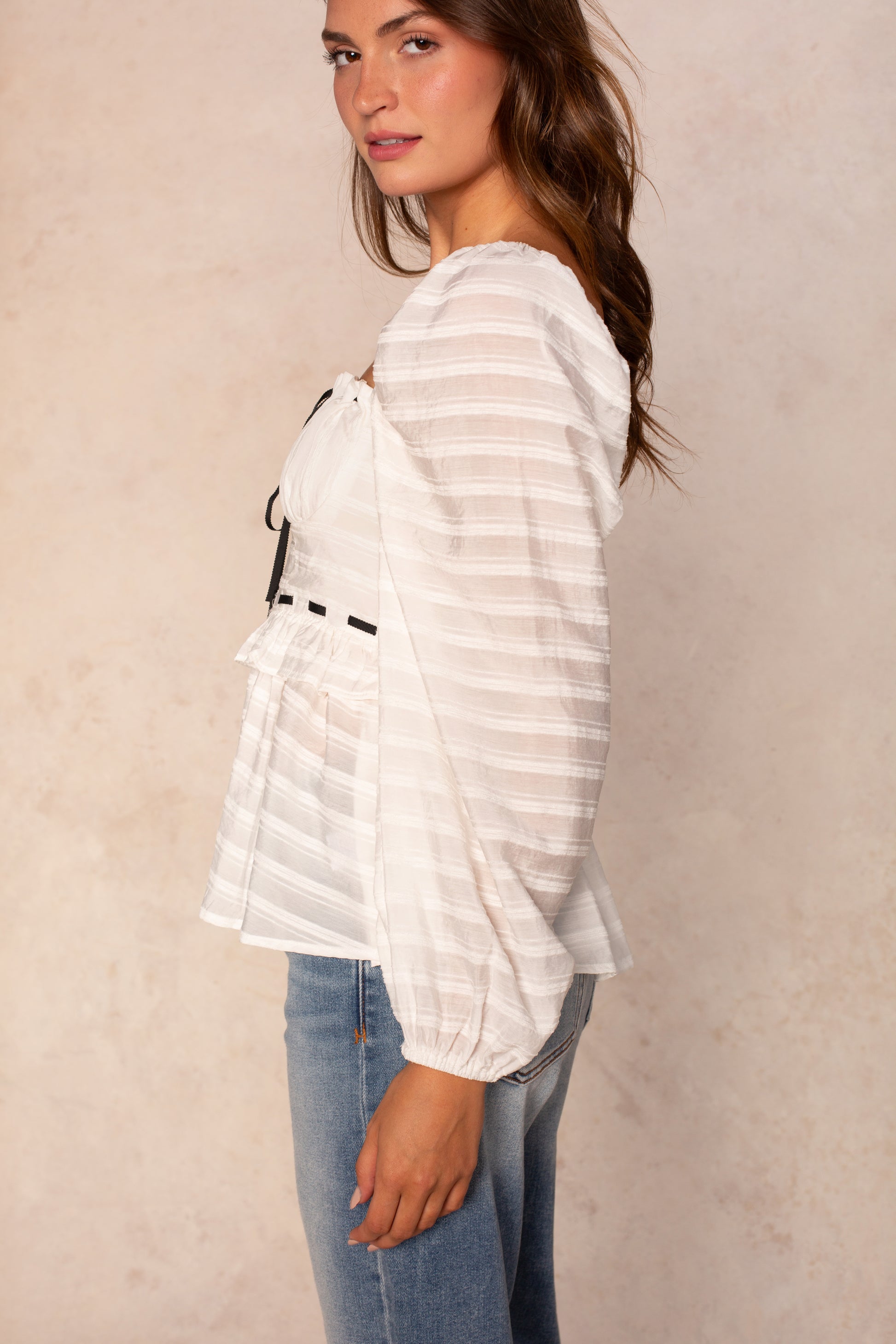 A woman with long brown hair stands sideways against a light beige background, wearing the Georgie Willow Longsleeve Blouse—a white, semi-sheer top with loose sleeves—paired with blue jeans.