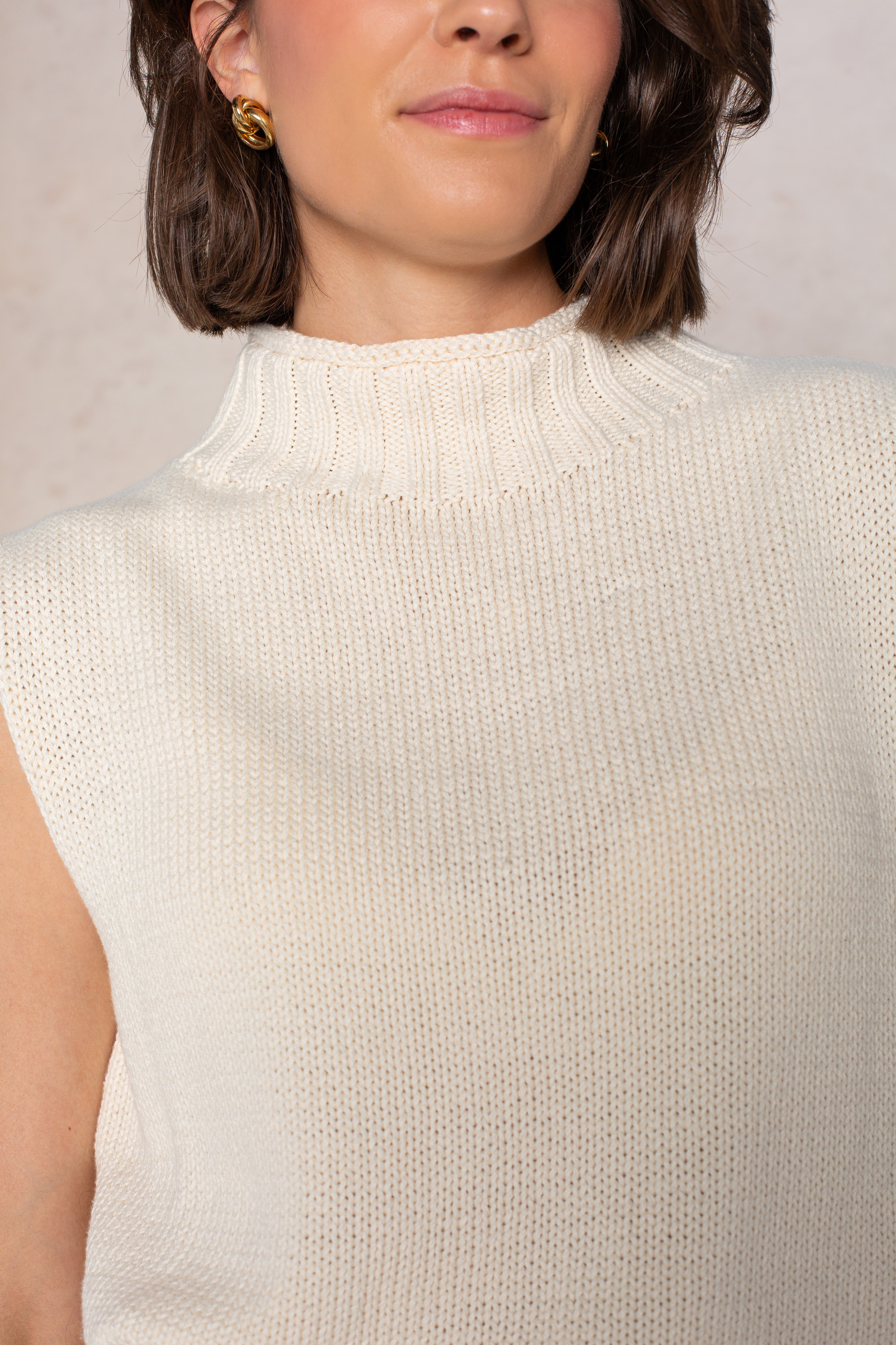 A woman with short brown hair wears gold hoop earrings and the Georgie Grace Rolled Neck Sleeveless Sweater in Ivory. The image, against a neutral background, is cropped to show her from the shoulders up.