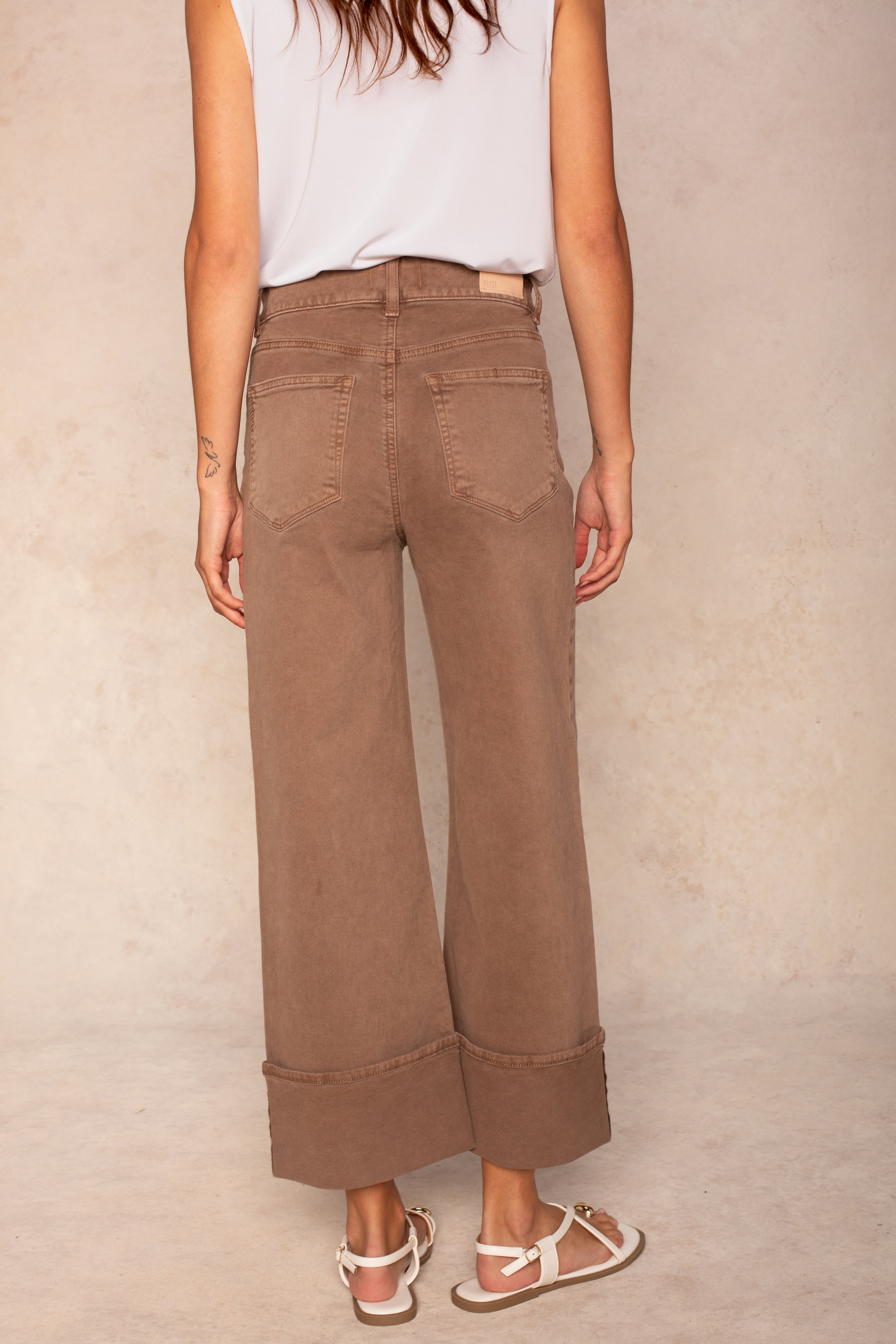 A woman stands facing away against a light beige background wearing the Georgie x Paige Sasha Cuff in Vintage Canewood, styled with a sleeveless white top and white sandals.