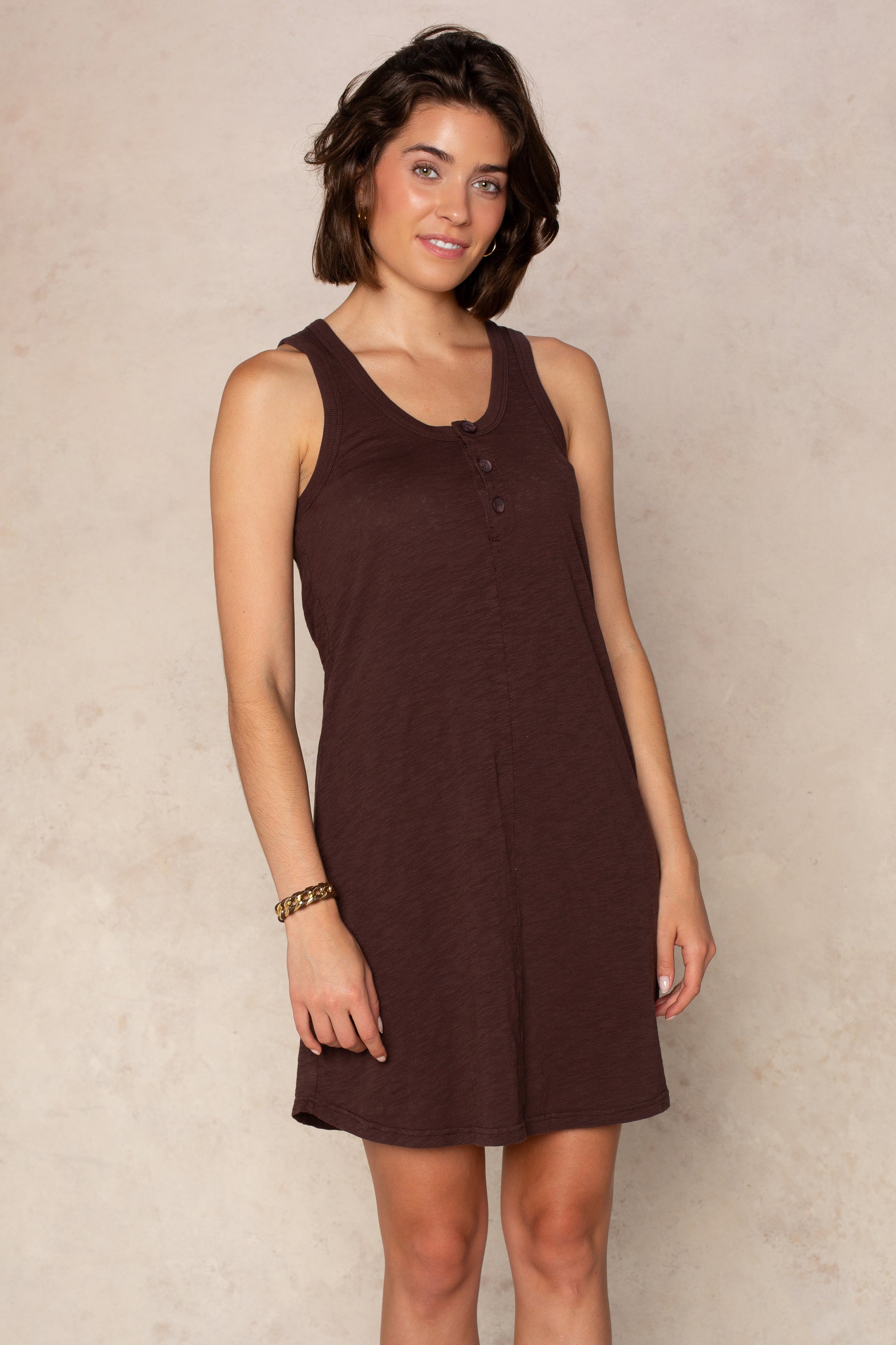 A woman with short brown hair smiles softly at the camera, wearing the Isla Zara Henley Tankdress Coverup—a sleeveless, brown, button-front mini dress—while standing against a light beige textured background.