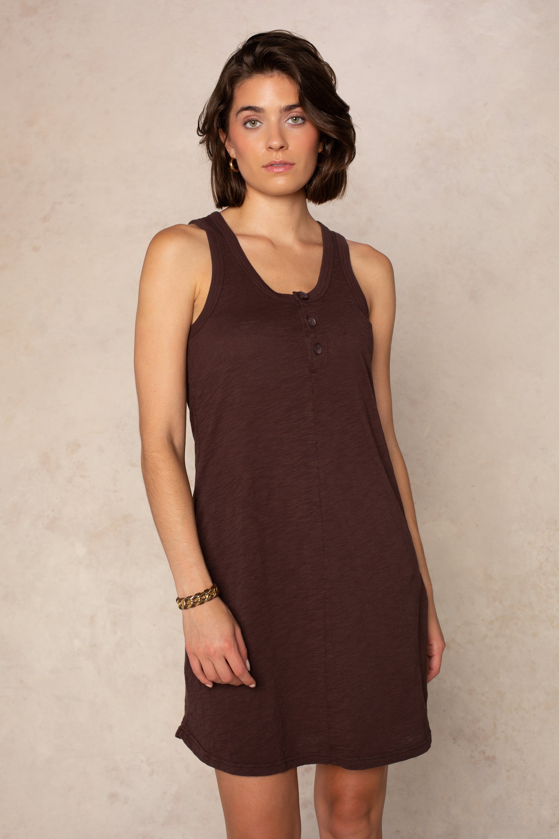 A woman with short brown hair and a neutral expression stands against a light textured background, wearing the Isla Zara Henley Tankdress Coverup in UMBER. The sleeveless, mini dress is perfect for layering over swim and accessorized with a gold bracelet.