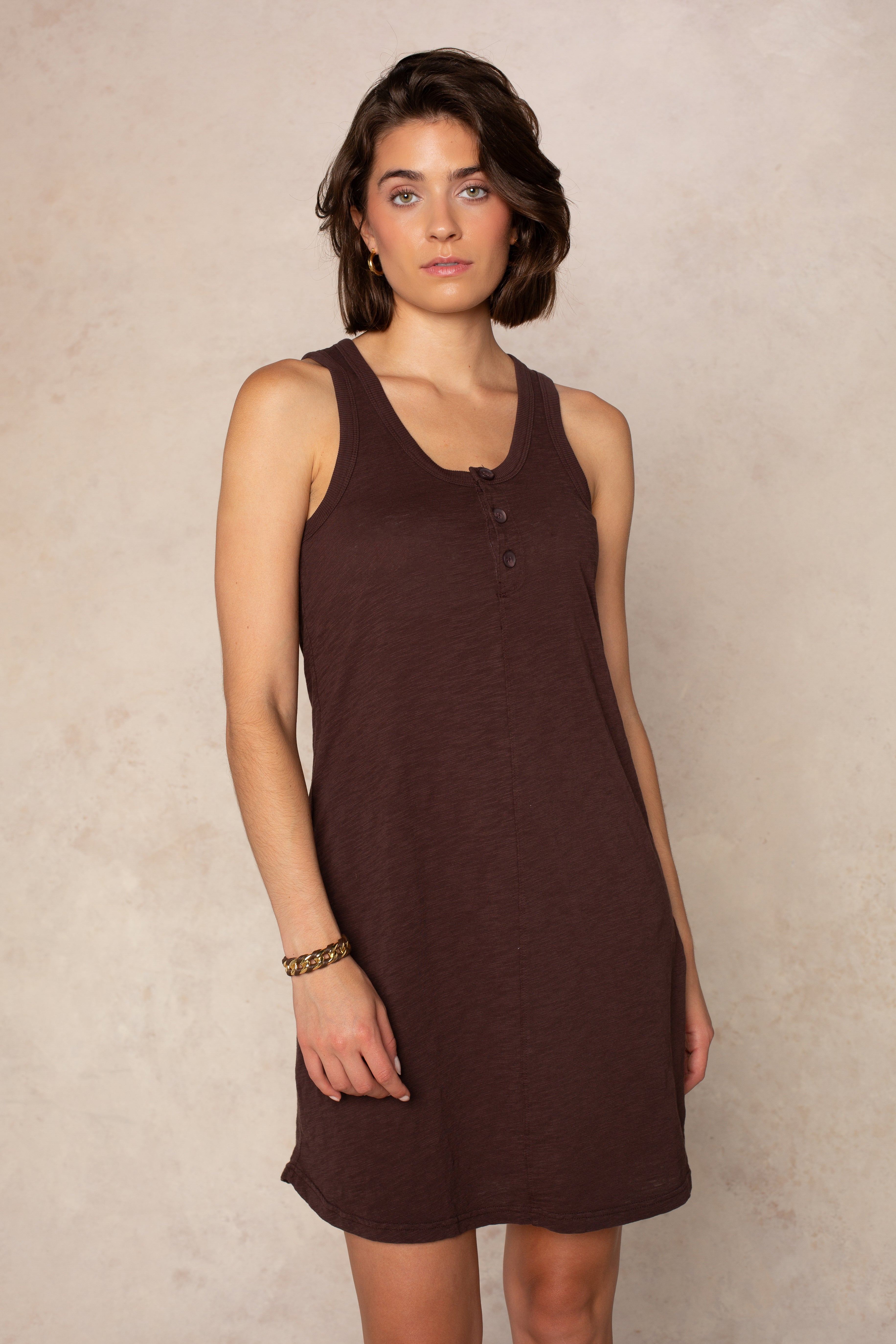 A woman with short brown hair and a neutral expression stands against a light textured background, wearing the Isla Zara Henley Tankdress Coverup in UMBER. The sleeveless, mini dress is perfect for layering over swim and accessorized with a gold bracelet.