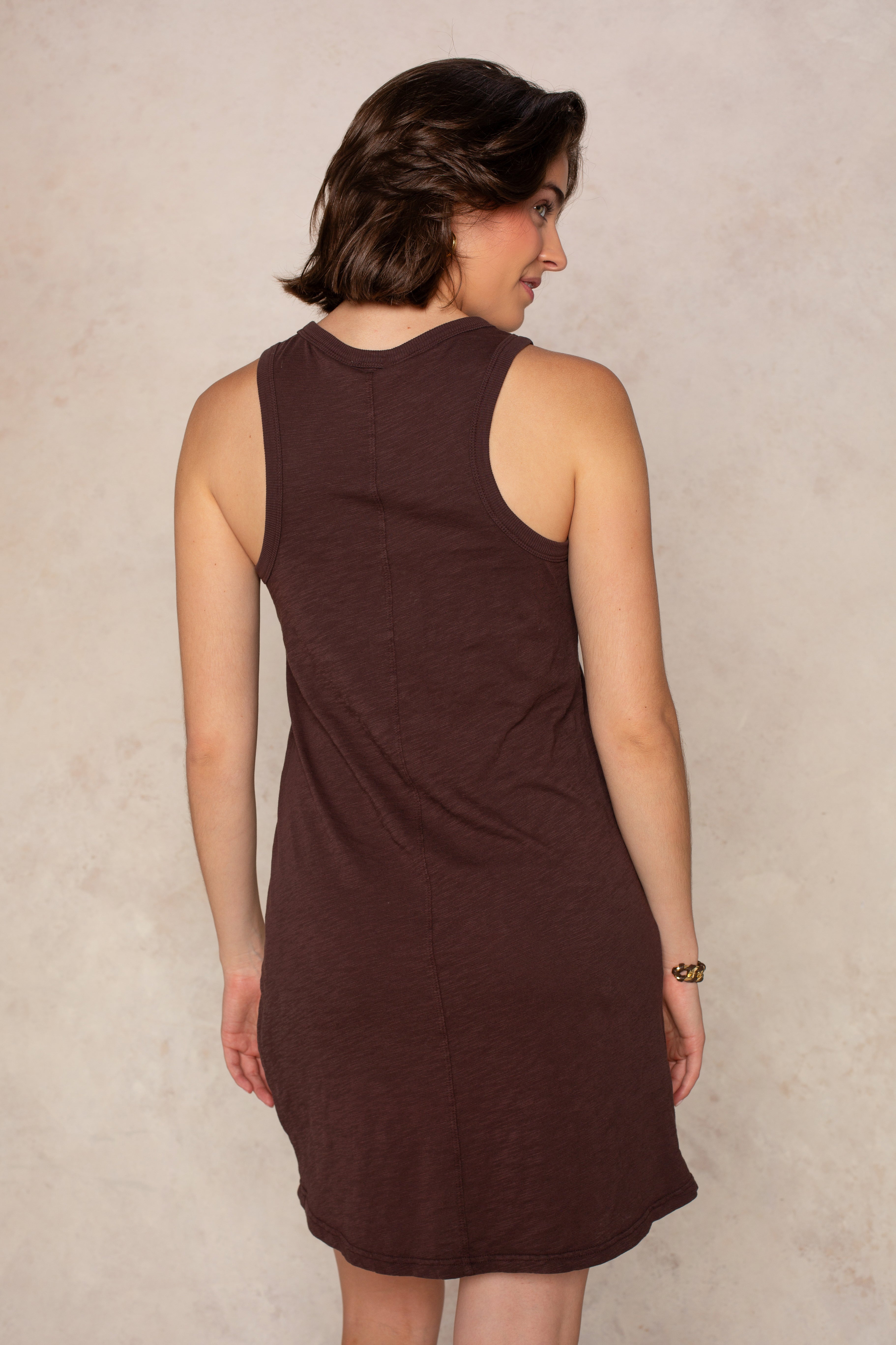 A woman with short brown hair faces away from the camera, wearing the Isla Zara Henley Tankdress Coverup in brown. The sleeveless dress is knee-length and set against a light, neutral wall background.