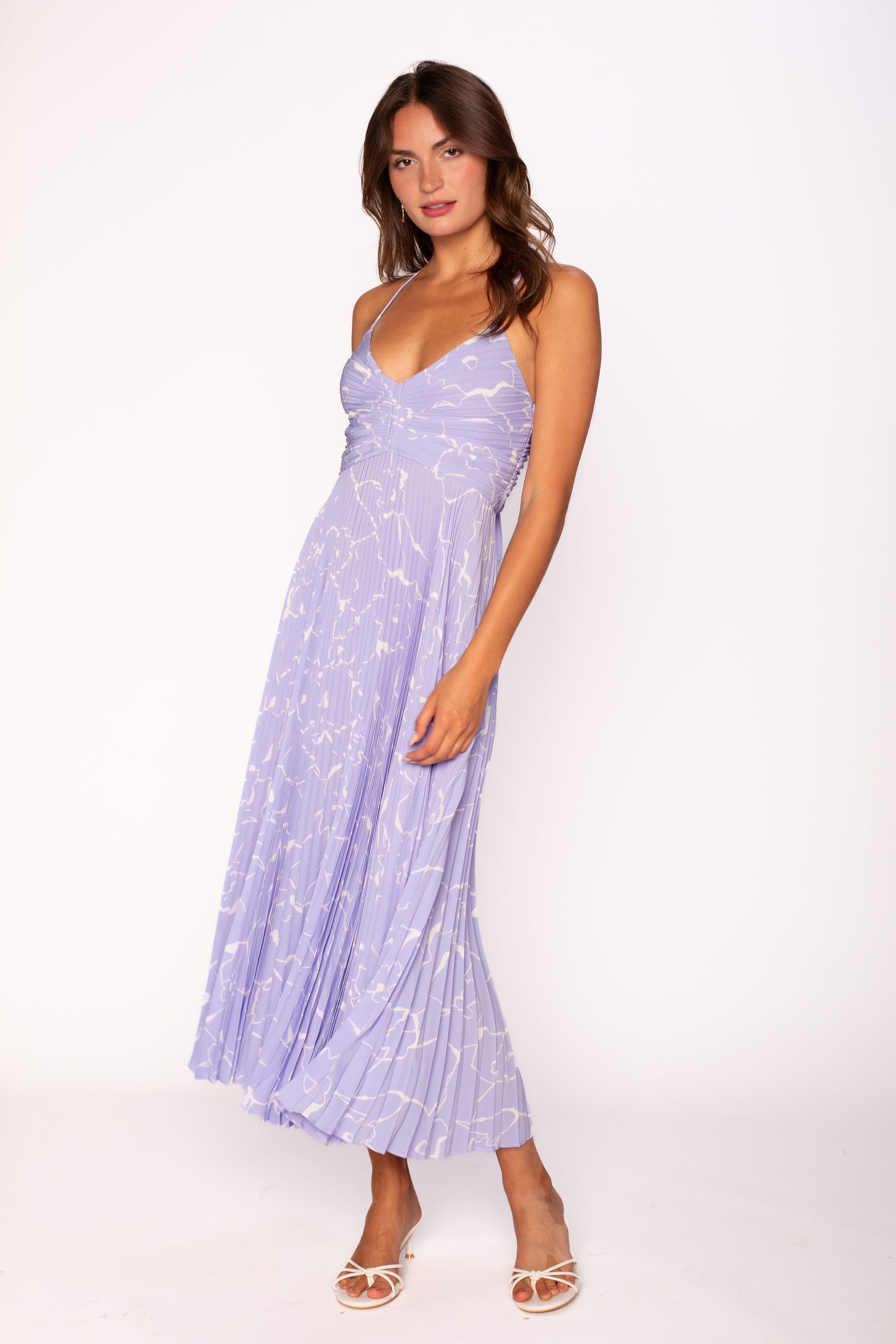 A woman with long brown hair wears the ASTR Blythe Midi Dress in Periwinkle Floral, featuring a light purple plissé fabric, V-neckline, and white floral pattern. She pairs it with white open-toe heels against a plain white background.