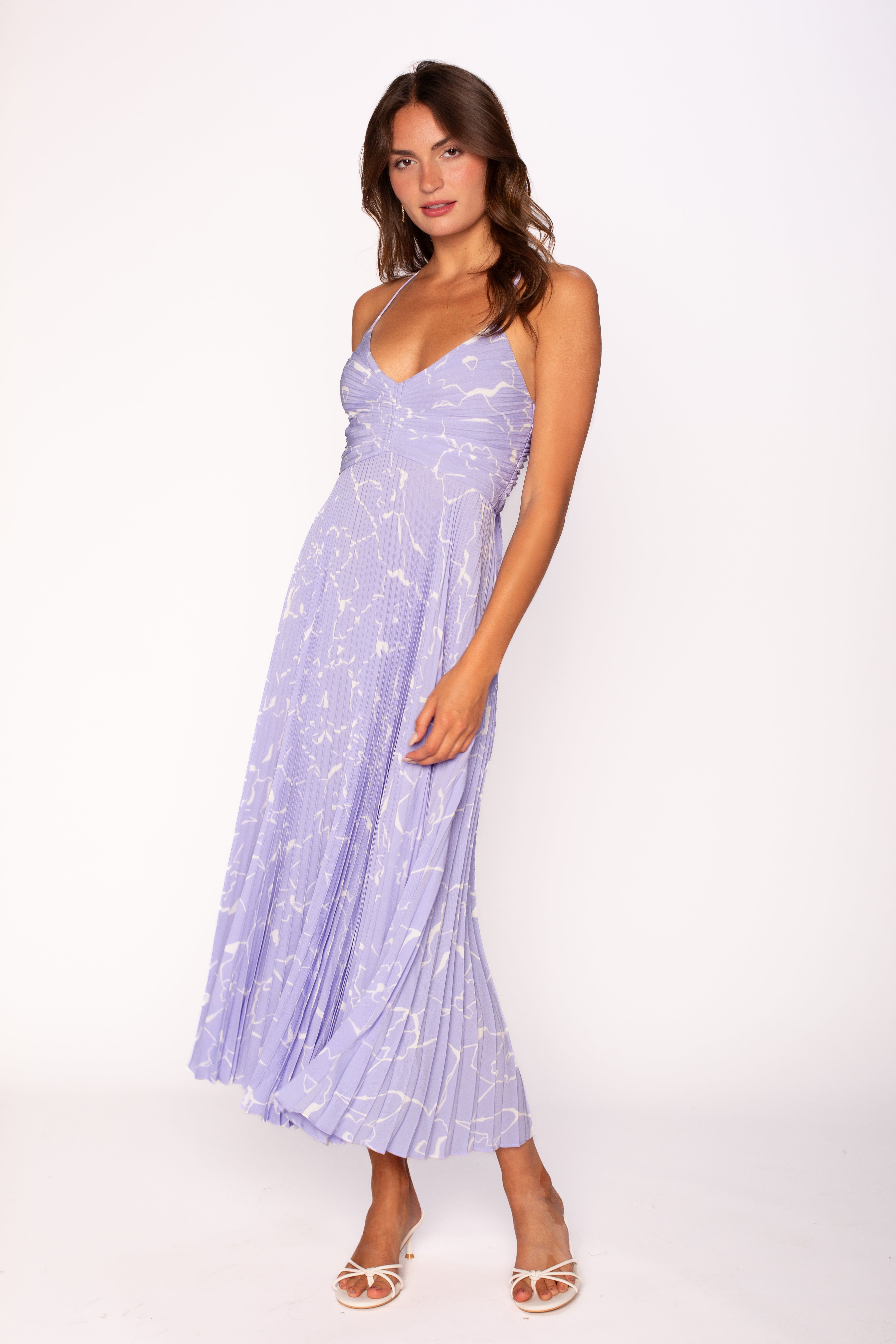 A woman with long brown hair wears the ASTR Blythe Midi Dress in Periwinkle Floral, featuring a light purple plissé fabric, V-neckline, and white floral pattern. She pairs it with white open-toe heels against a plain white background.