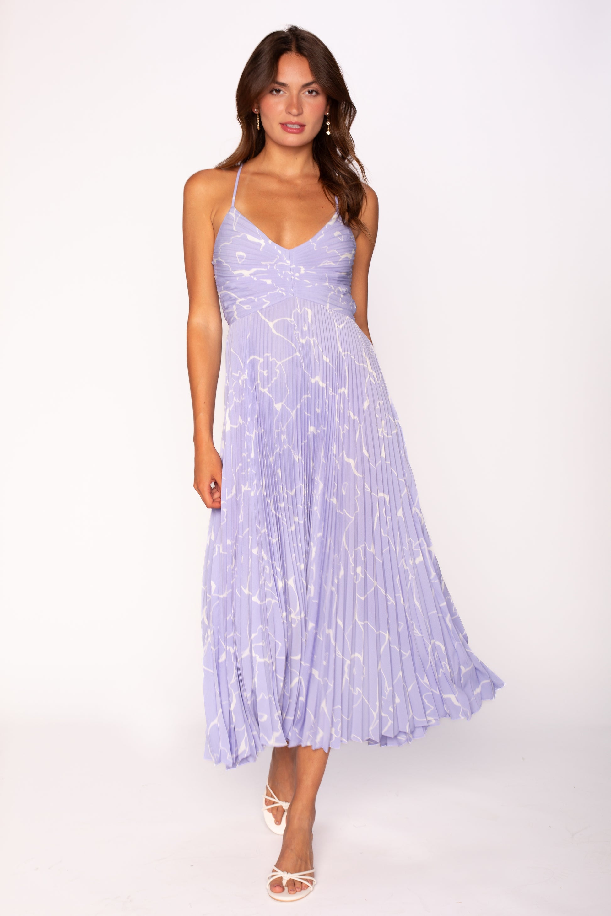 A woman stands against a white background wearing the Blythe Midi Dress by ASTR in light blue, featuring adjustable spaghetti straps and a white abstract pattern, paired with white open-toe heels.