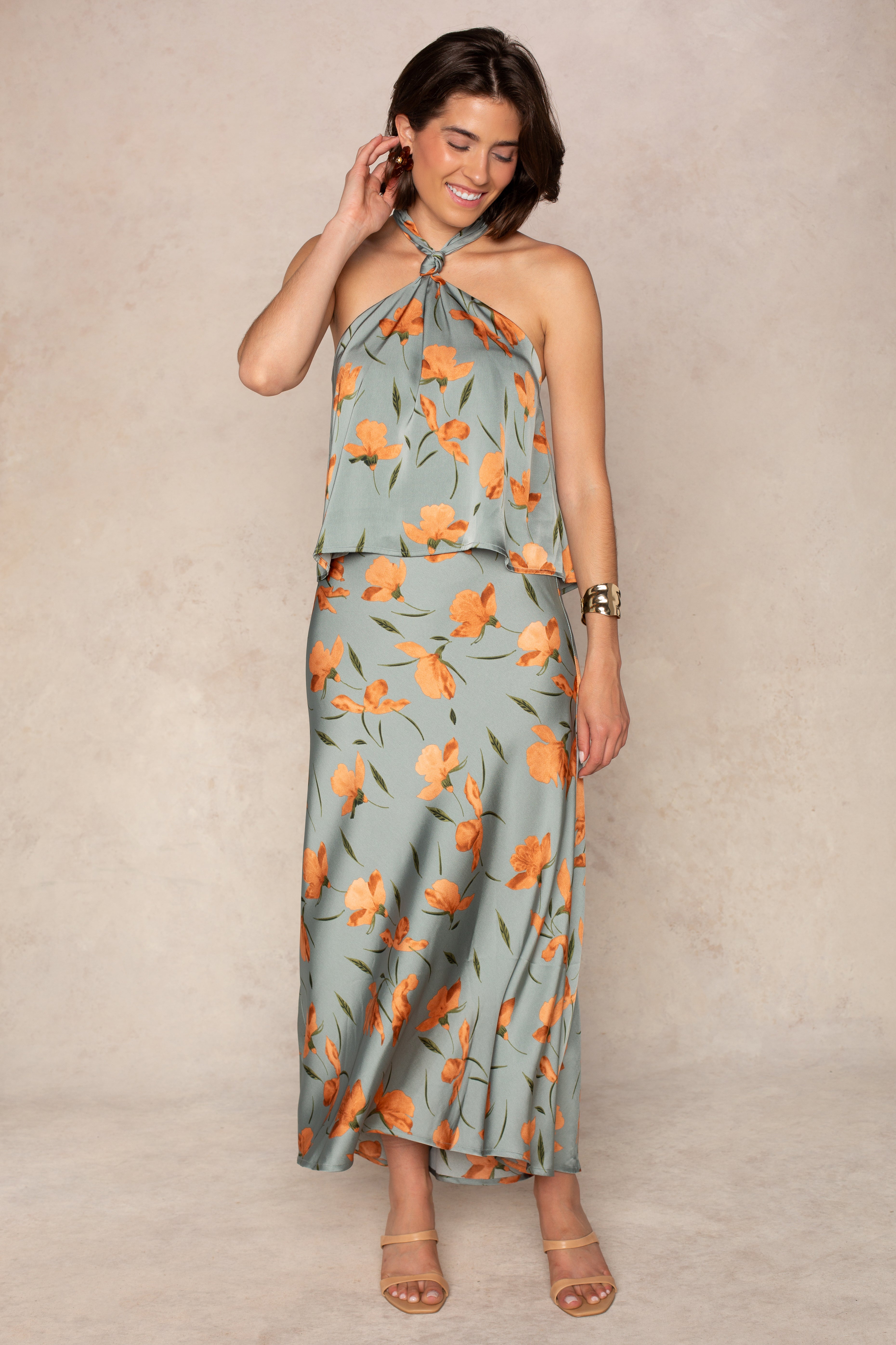 A woman smiles, wearing the Georgie Emilia Satin Skirt—a light blue piece with an orange floral print—paired with nude heels, hoop earrings, and a bracelet. One hand touches her hair.
