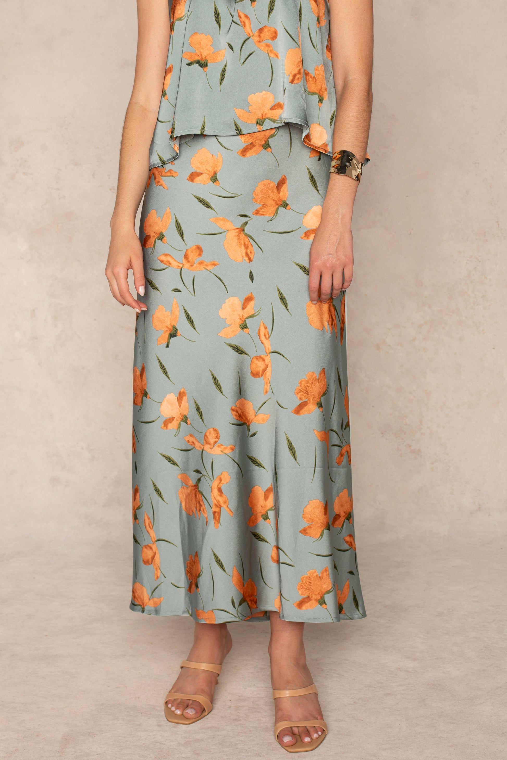 A person stands against a neutral background wearing a light green sleeveless top and the Georgie Emilia Satin Skirt in sage with an orange floral print, paired with nude sandals and a gold bracelet.