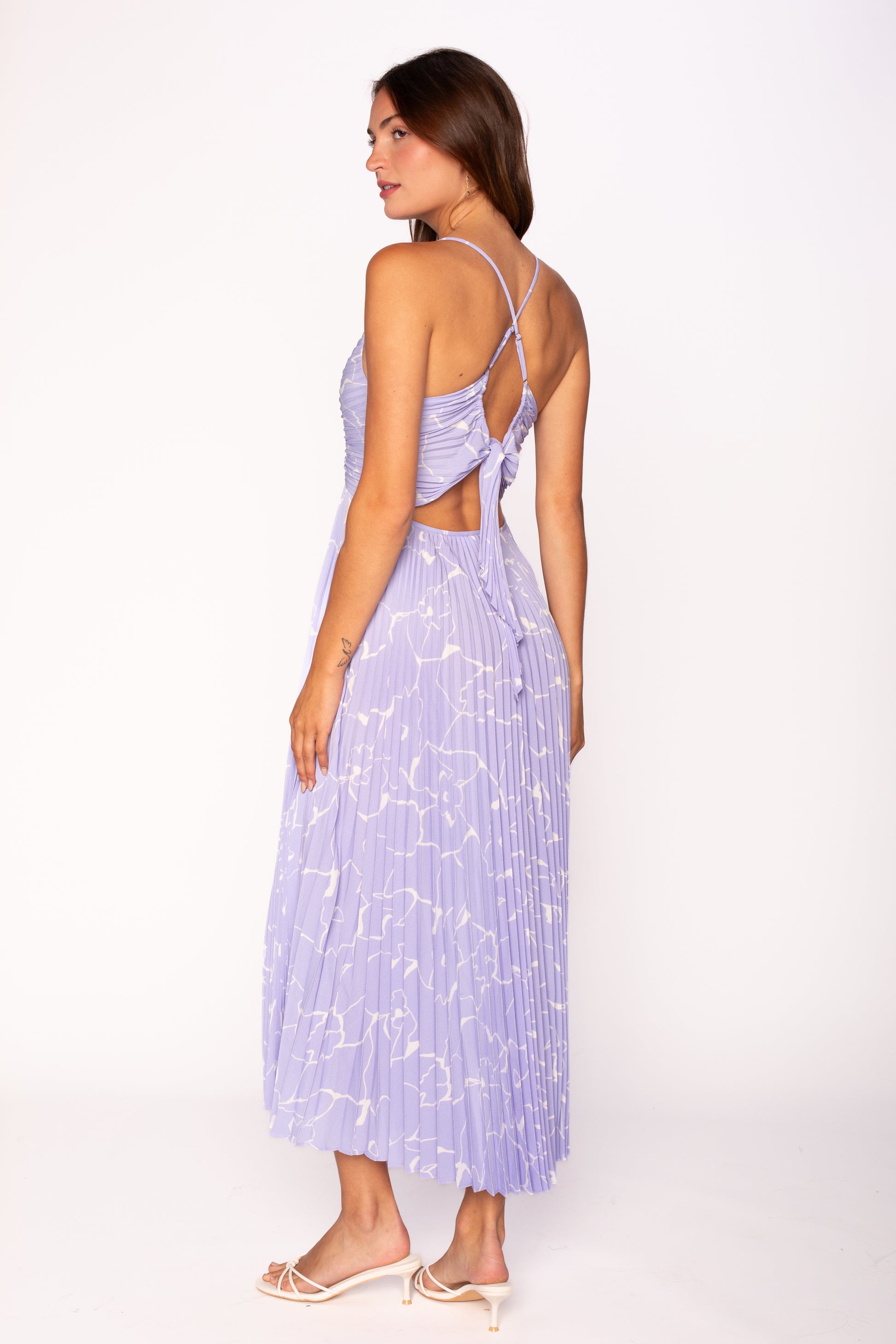 A woman models the ASTR Blythe Midi Dress—a light purple plissé dress with white abstract patterns, open back with crisscross adjustable spaghetti straps—paired with white heels, standing slightly turned on a white background.