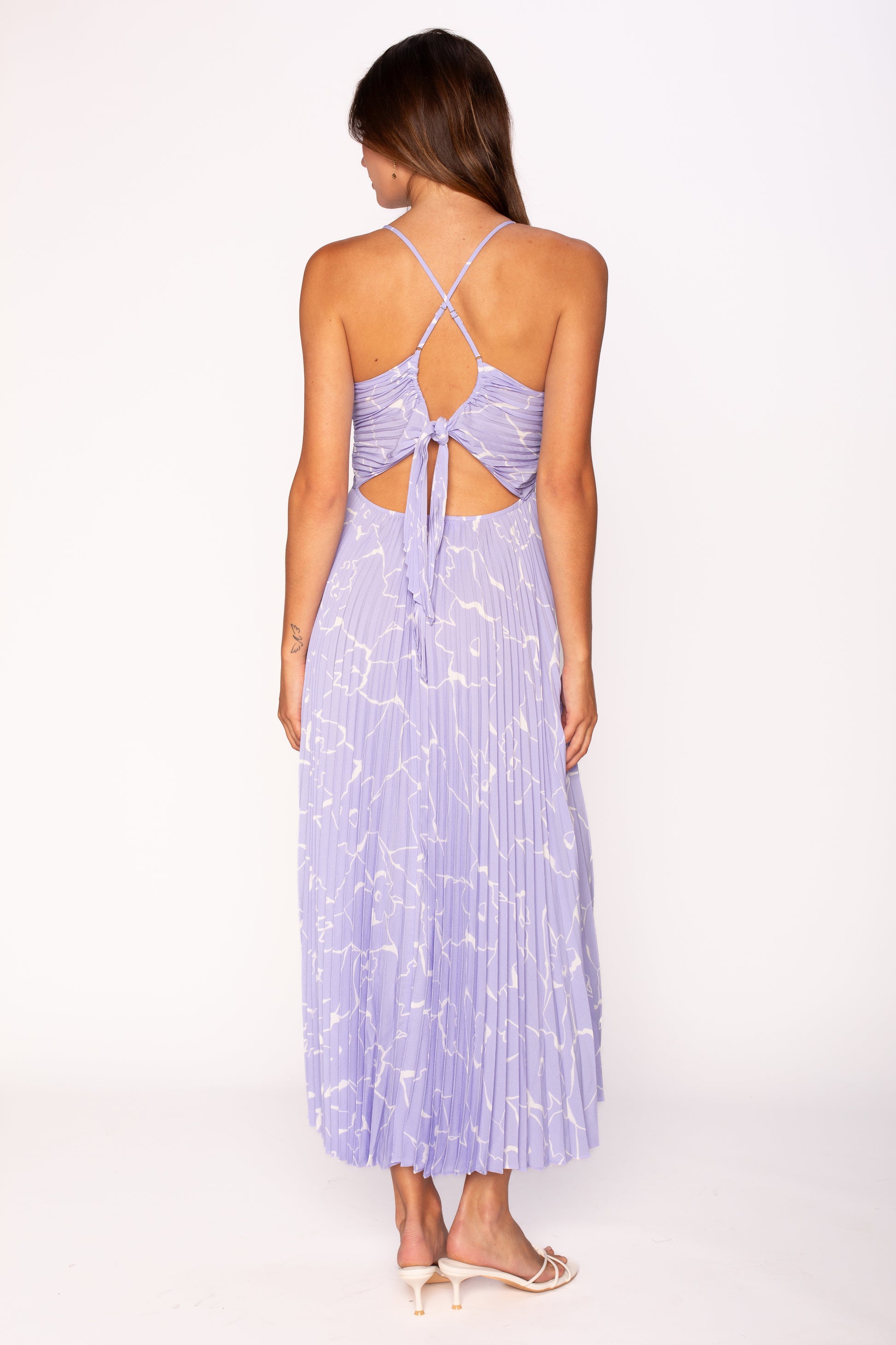 A woman faces away, wearing the ASTR Blythe Midi Dress in light purple with white patterns, featuring an open back, adjustable crisscross spaghetti straps, and a bow tie. She pairs it with light heeled sandals.
